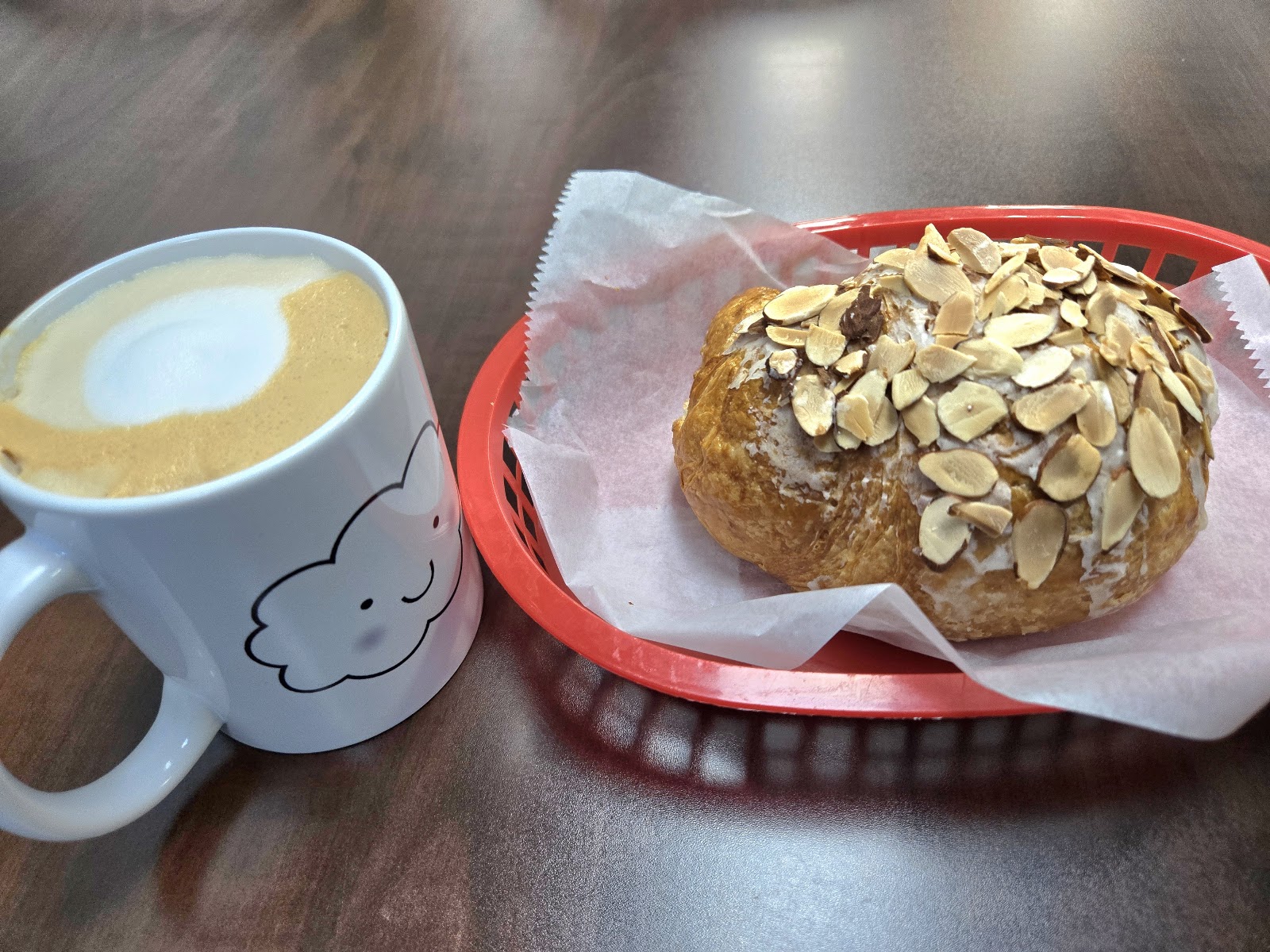 A glazed almond croissant topped with sliced almonds in a basket, next to a latte in a white mug printed with the Cloud Cafe smiling-cloud logo