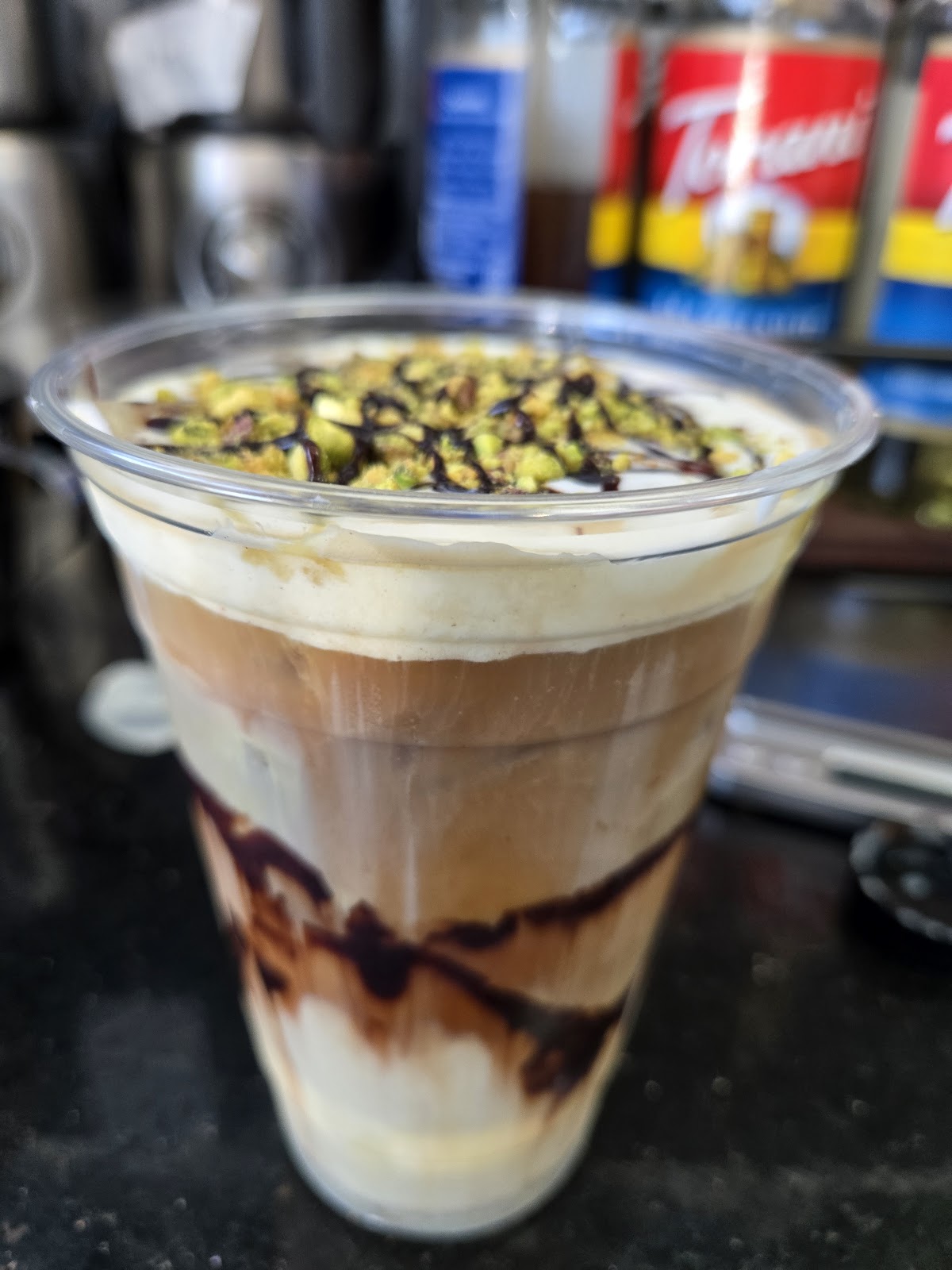 A tall iced layered latte with cream on top, a chocolate drizzle, and crushed pistachio, in a clear cup
