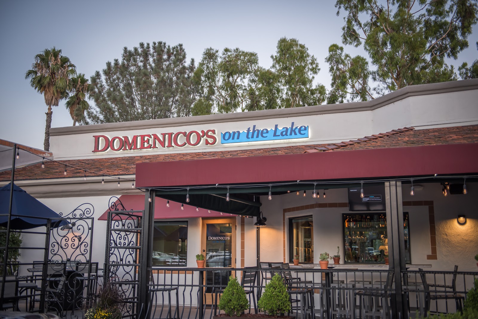 The Domenico's on the Lake storefront at dusk, palm trees and a patio with iron chairs, the sign glowing
