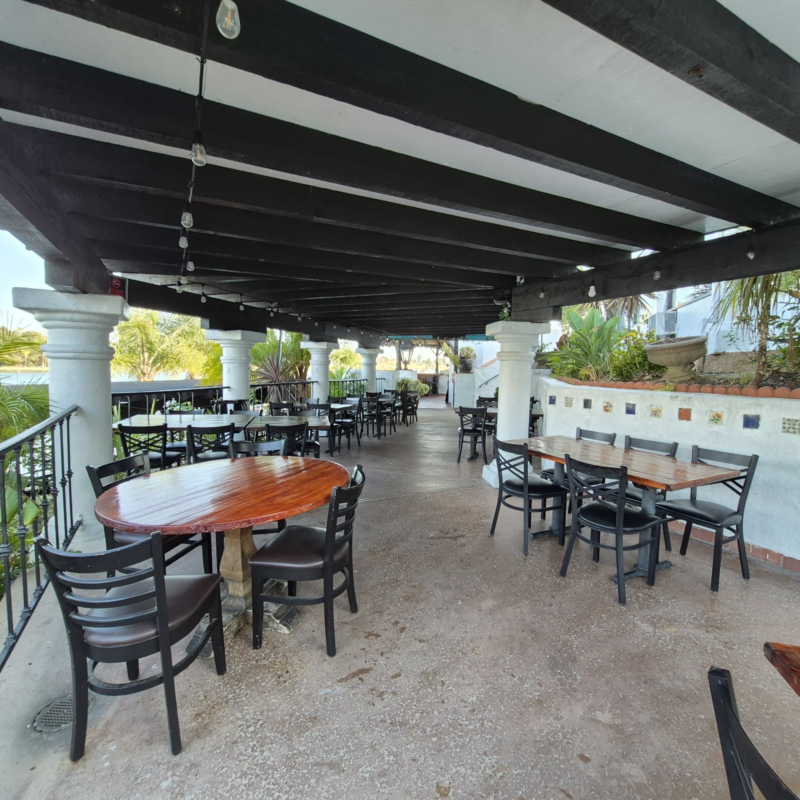 The lakeside terrace at Domenico's on the Lake. Long covered patio with iron-frame chairs and wooden tables, sun streaming through the pillars, the lake visible past the railing.