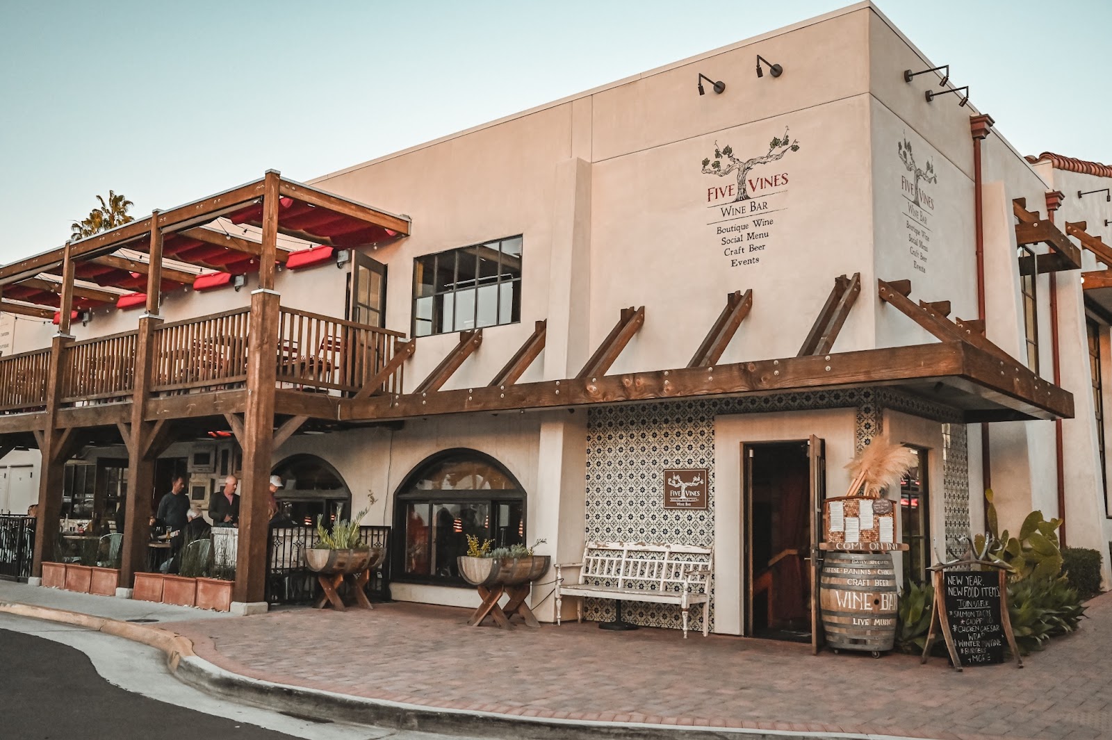 The Mission-style two-story Five Vines Wine Bar building in San Juan Capistrano at dusk, with red awnings and a wine-bar chalkboard out front.