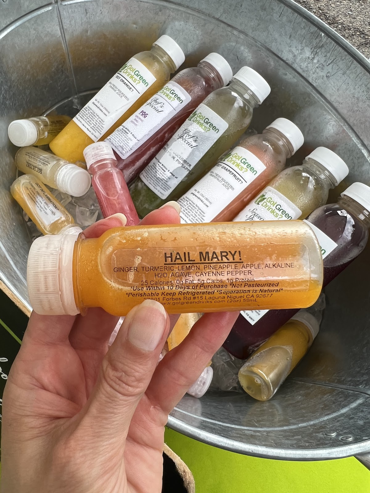 A hand holding a bright orange Hail Mary wellness shot over a galvanized tub of chilled Got Green Drinks juices on ice