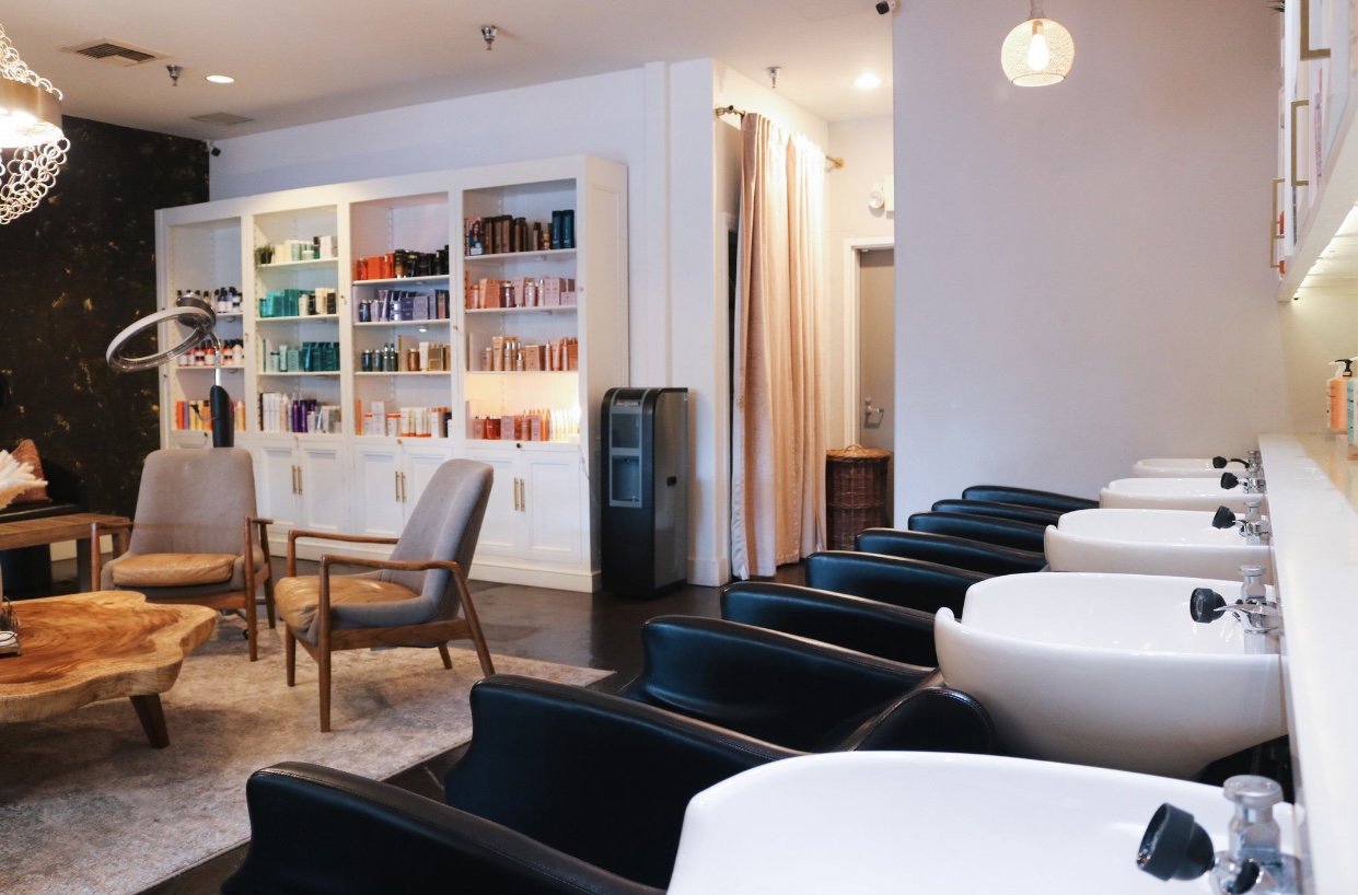 Row of wash stations with black basins and chairs, white product cabinets, and mid-century lounge seating inside H Studio Salon