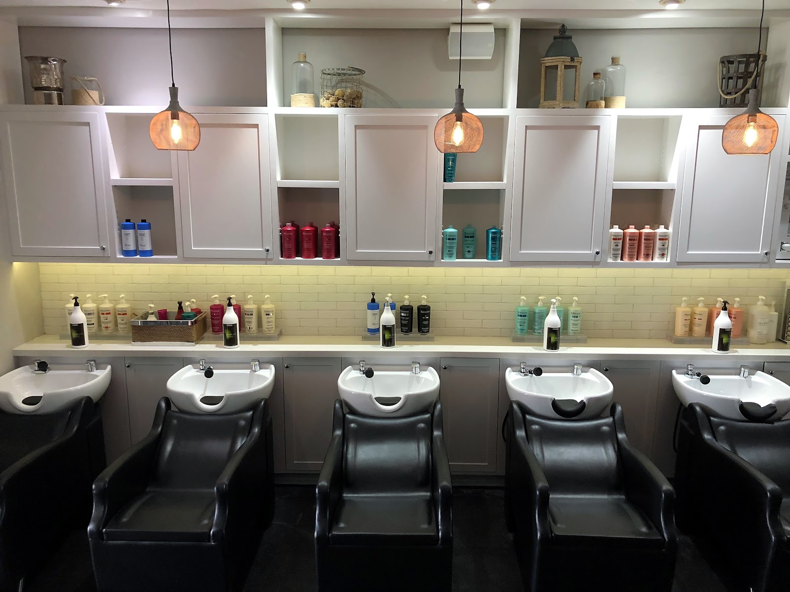 A second row of wash stations with subway-tile backsplash, copper pendant lights, and shelves of professional color products