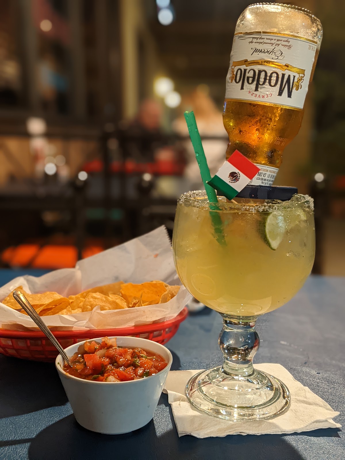 A basket of tortilla chips with a bowl of fresh red salsa beside a margarita topped with an inverted Modelo and a small Mexican flag at La Cocina de Ricardo