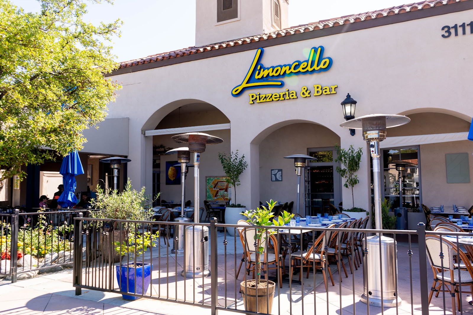 The sunlit terrace at Limoncello, blue umbrellas and bistro tables under arched stucco, the yellow Limoncello Pizzeria & Bar sign above