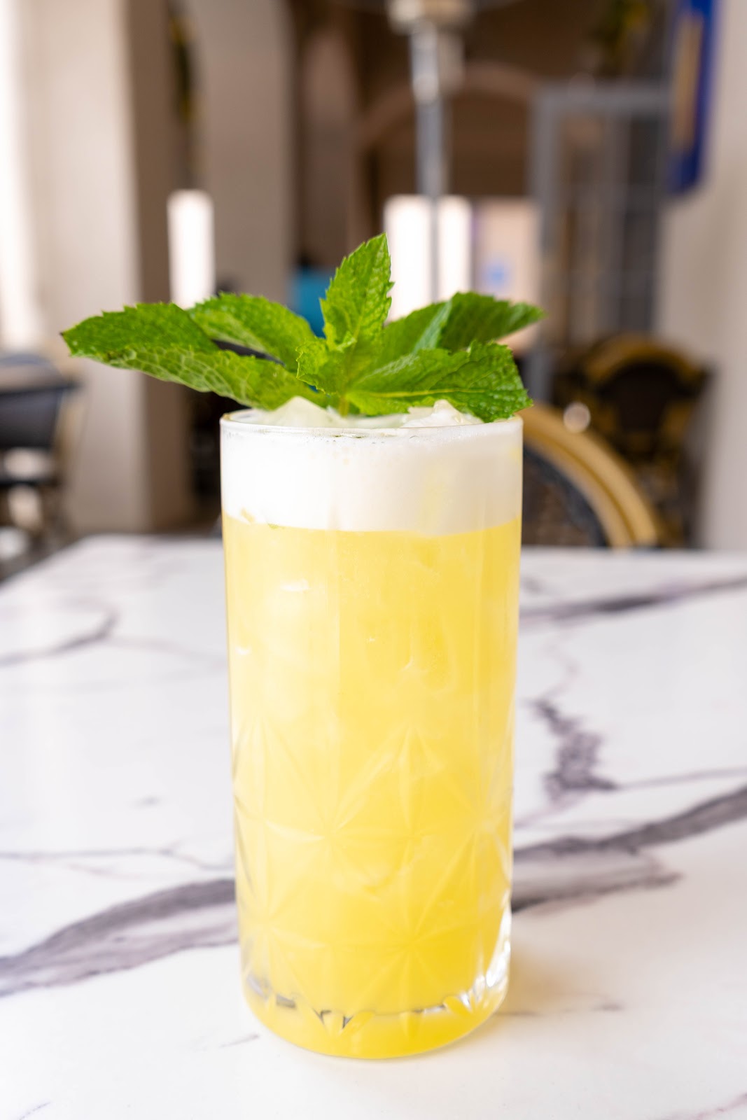 A frothy lemon-yellow gin sour with a sprig of mint in a faceted highball on a marble bar