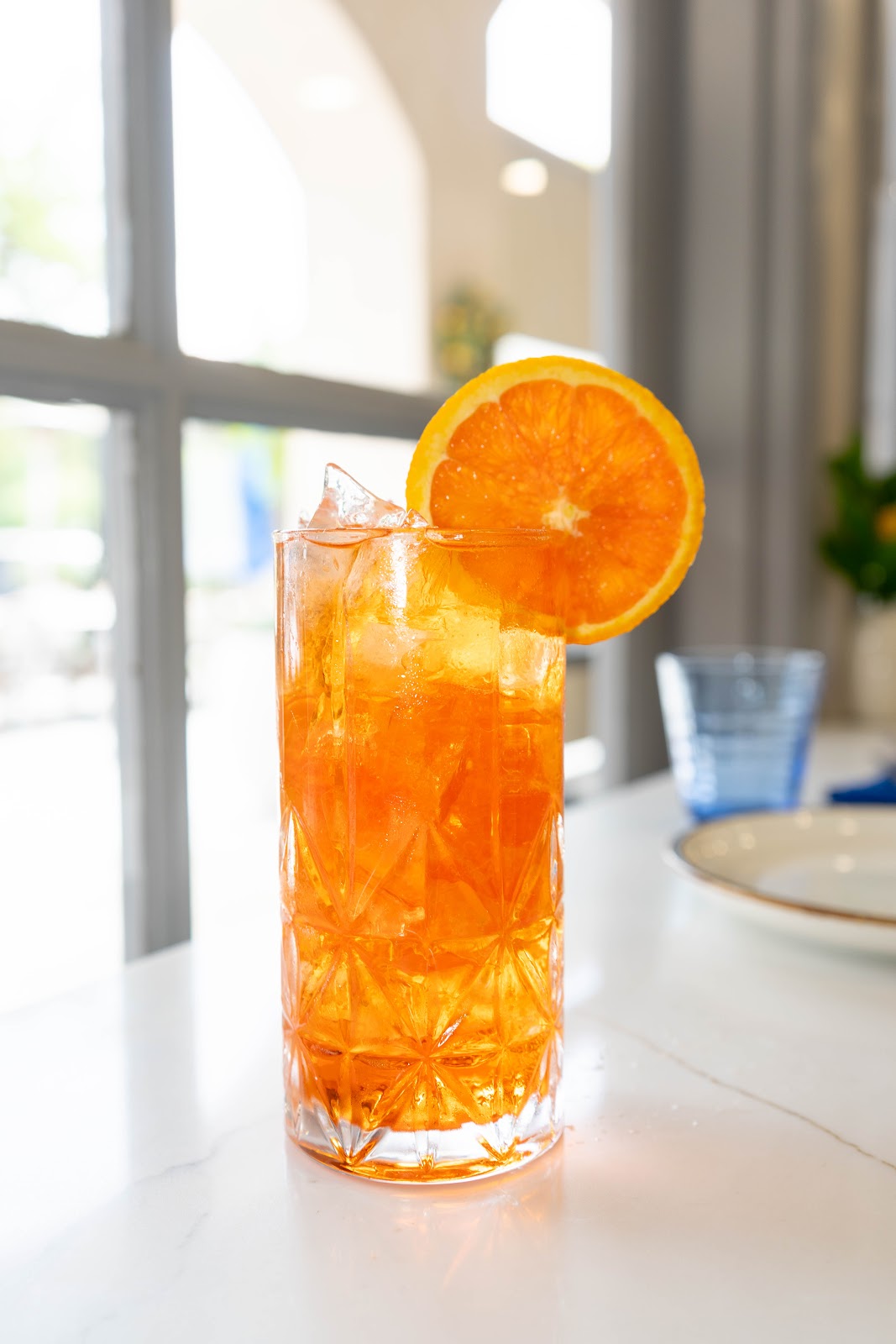An orange Aperol spritz over ice with an orange wheel, set by an arched window full of afternoon light