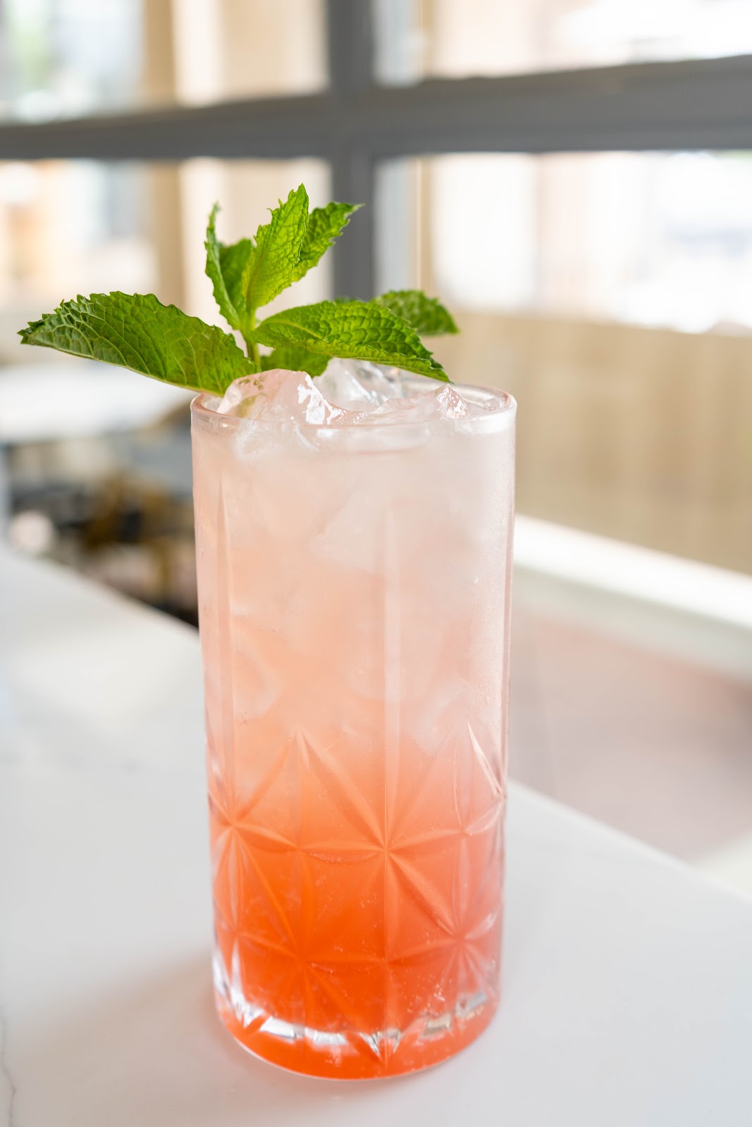 A pink strawberry mule in a tall faceted glass with crushed ice and a sprig of mint by a sunlit window