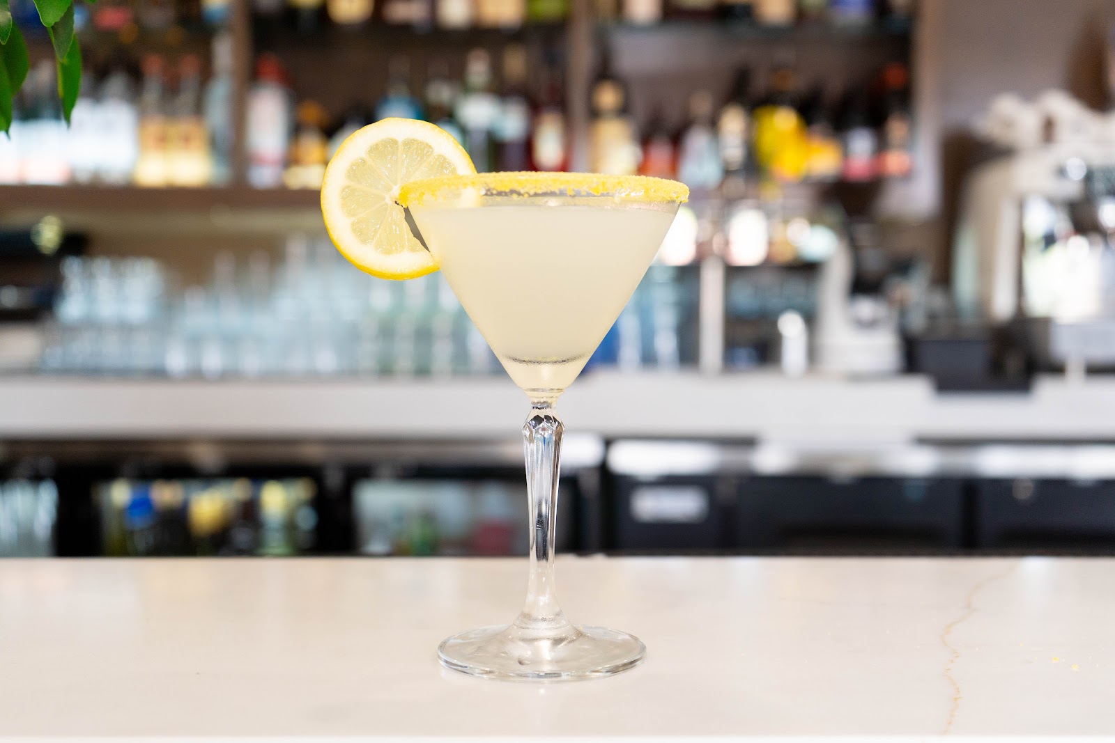 A lemon drop martini with a sugared rim and a lemon wheel on a marble bar in front of the back bar bottles