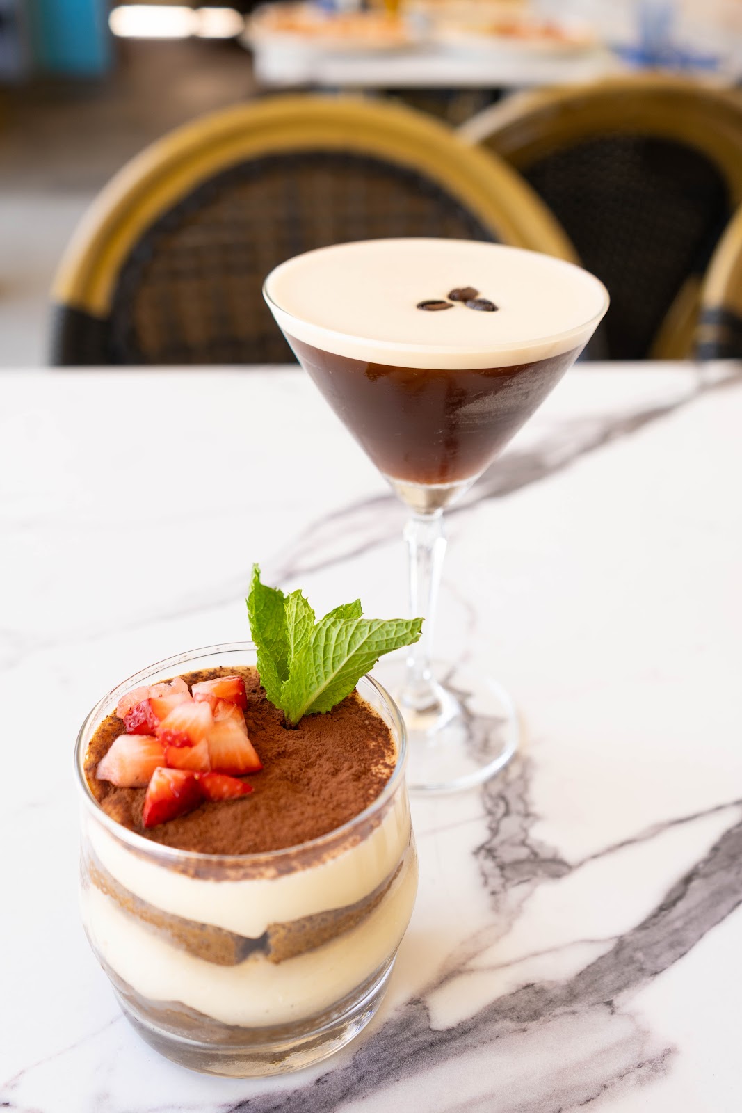 Tiramisu layered in a glass dusted with cocoa and topped with strawberries and mint, an espresso martini behind it on marble