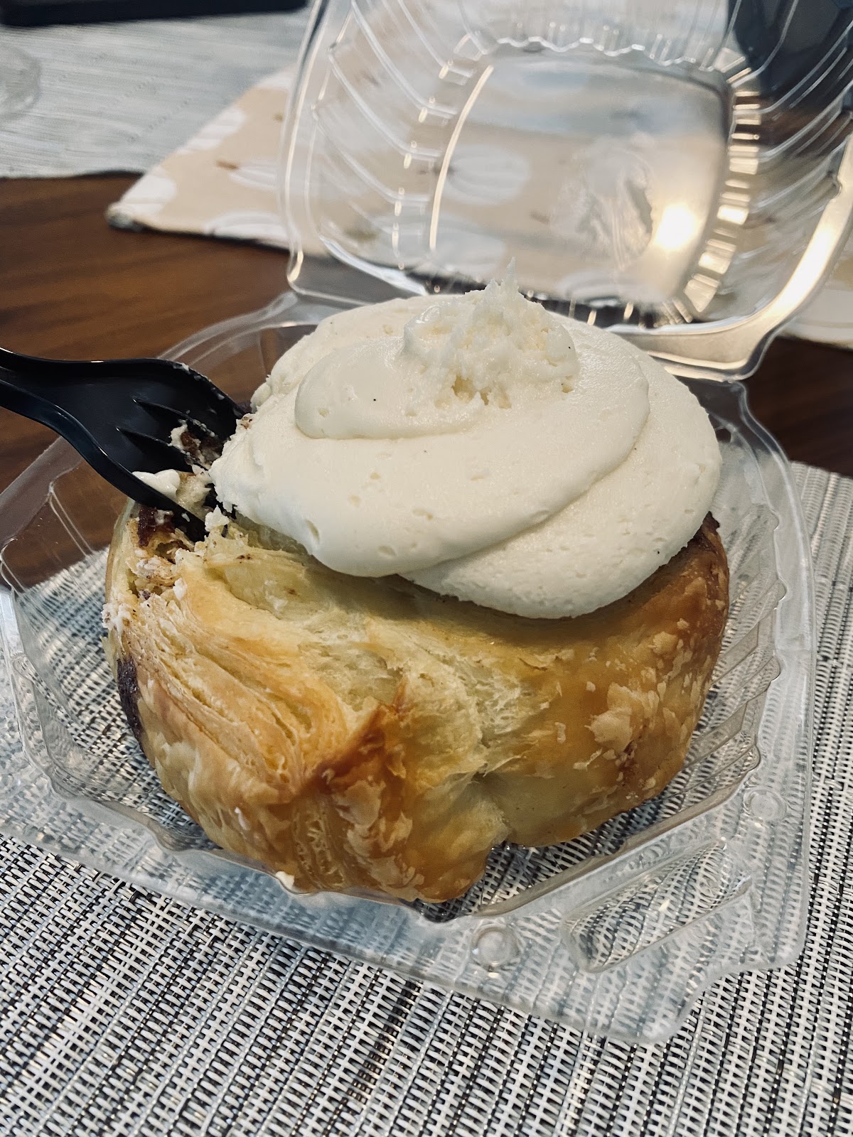 A cinnamon roll topped with a thick swirl of vanilla cream in a clear clamshell