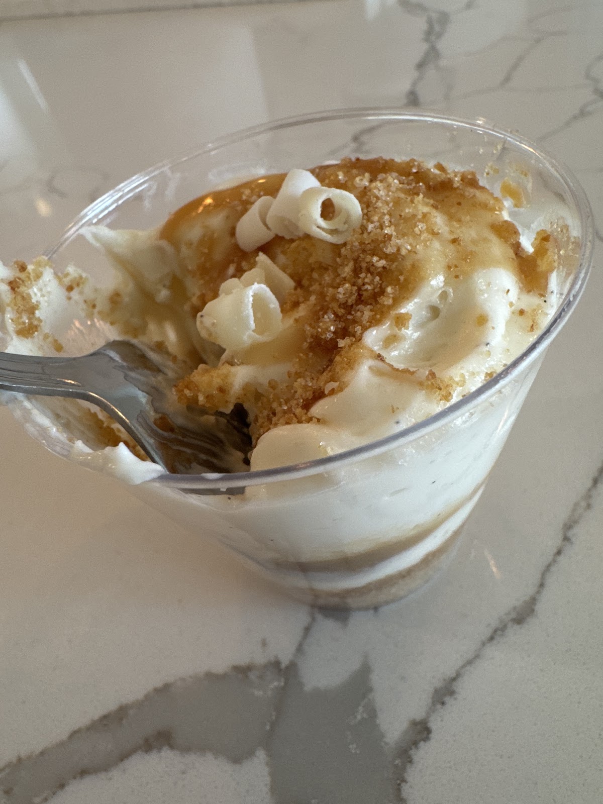 A caramel cheesecake cup topped with white chocolate curls and a graham crumble on a marble counter