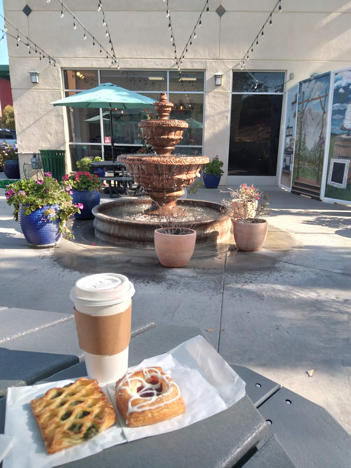 A danish and a savory croissant with a coffee on a patio table beside a stone fountain at A Little Something Sweet
