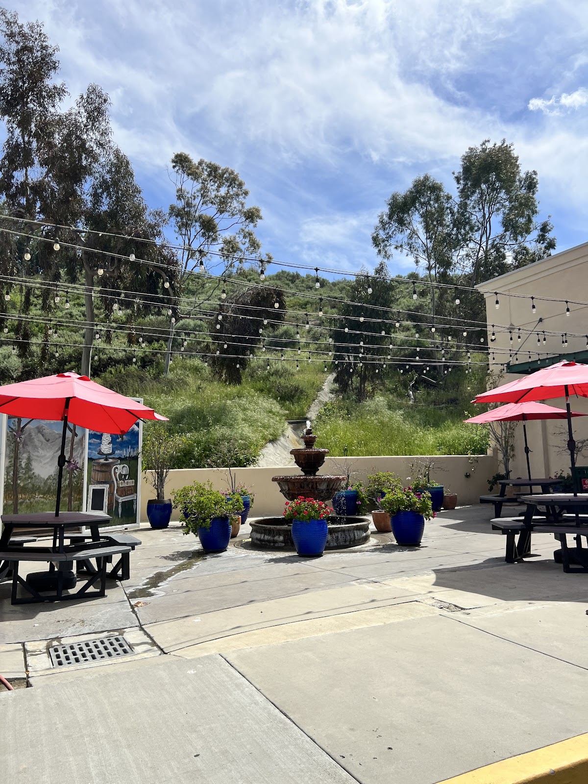 The outdoor patio at A Little Something Sweet with red umbrellas, a stone fountain, string lights and a green eucalyptus hillside behind