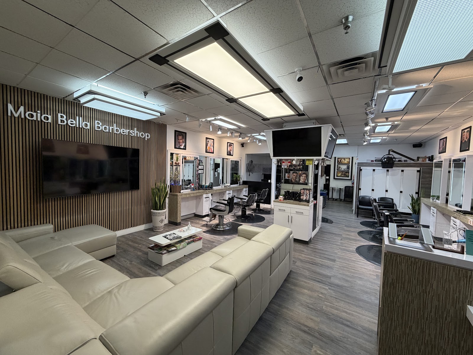 The bright modern interior of Maia Bella Barber and Salon in Lake Forest, with a cream leather sectional couch in the front lounge, a slatted wood feature wall carrying the Maia Bella Barbershop wall sign, and the barber stations and mirrors beyond