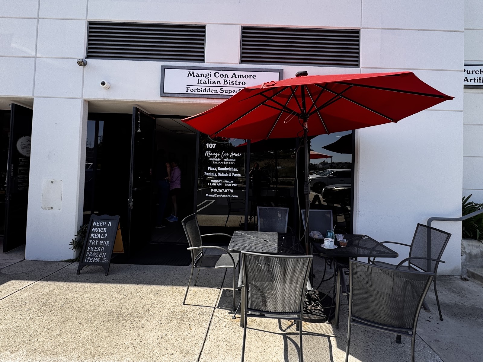 The storefront of Mangi Con Amore Italian Bistro at 107 Cabot Road, with the red patio umbrella, patio chairs, and the chalk sign reading TRY OUR FRESH FROZEN ITEMS