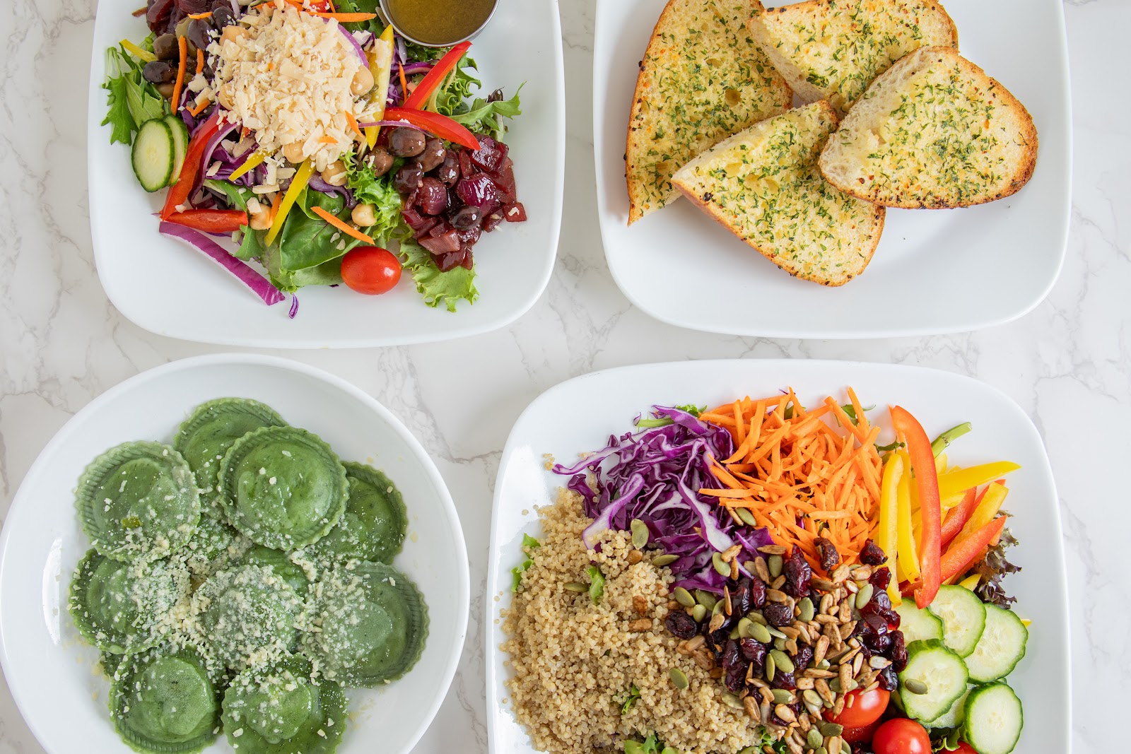 A table spread at Mangi Con Amore: a Mediterranean salad, garlic bread, spinach ravioli with parmigiano, and a quinoa veggie bowl with cabbage and seeds