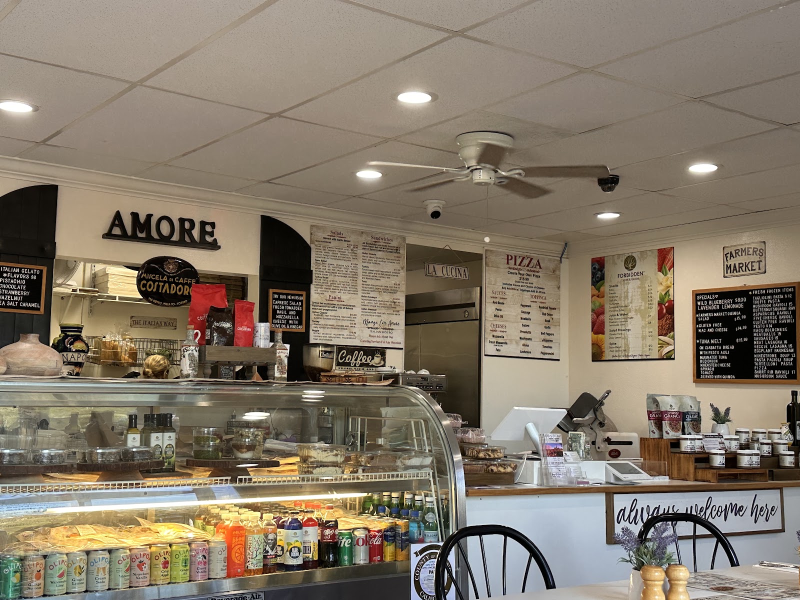 The counter at Mangi Con Amore with the AMORE sign on the wall, Costadoro espresso bags on the counter, hand-written menu boards for Salads, Panini, Pizza and Forbidden Superfoods, and the always-welcome-here sign by the dining tables