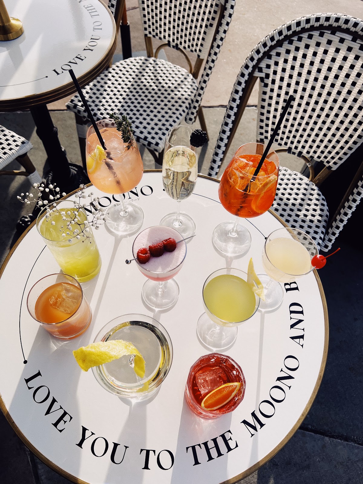 A round bistro table on the patio at MOONO with a flight of cocktails, the wordmark LOVE YOU TO THE MOONO AND BACK in serif type wrapping the table rim