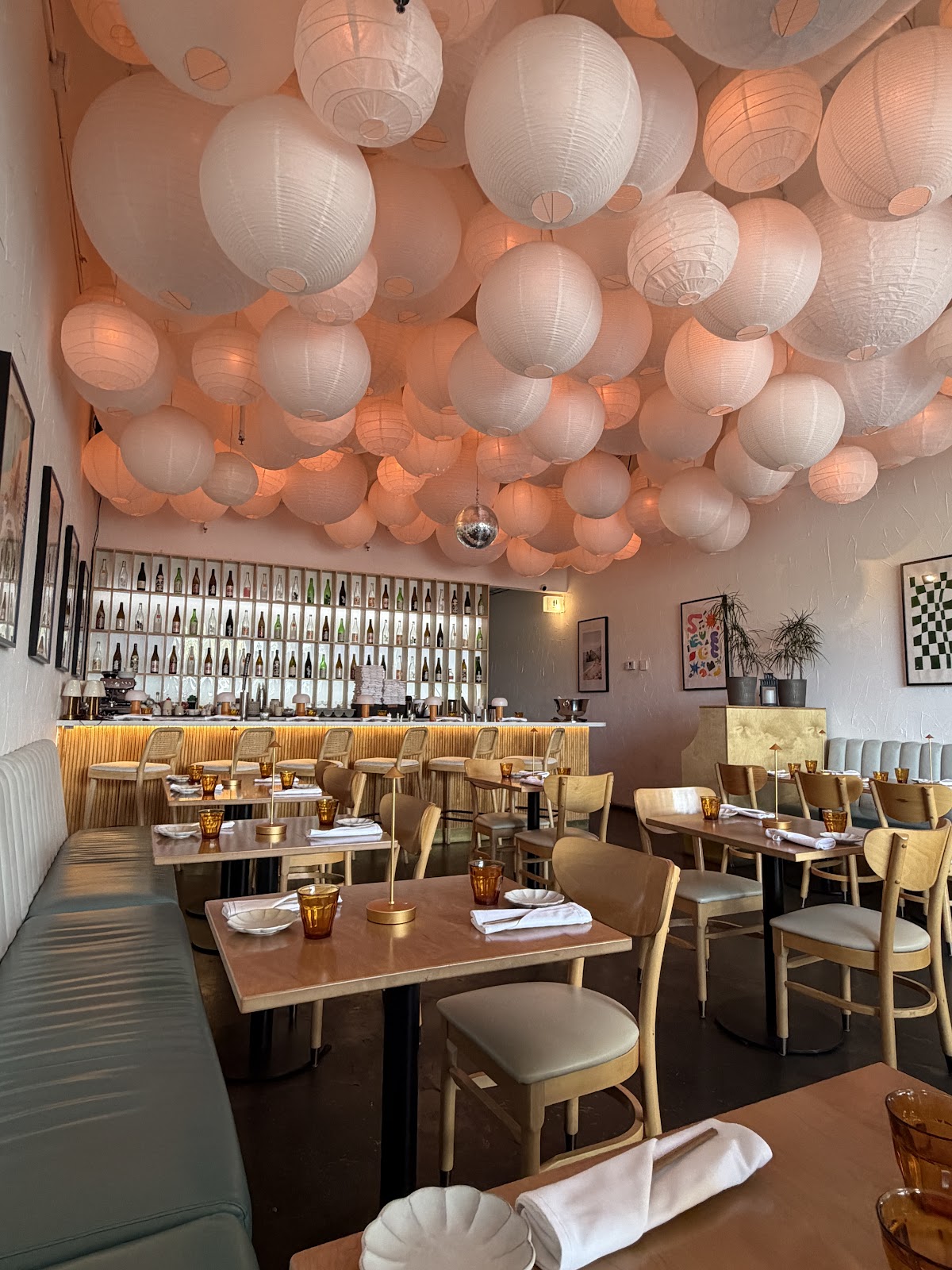 The dining room at MOONO: a ceiling clustered with white paper chochin lanterns over blonde wood tables, a sake-bottle wall behind the bar, green leather banquette below