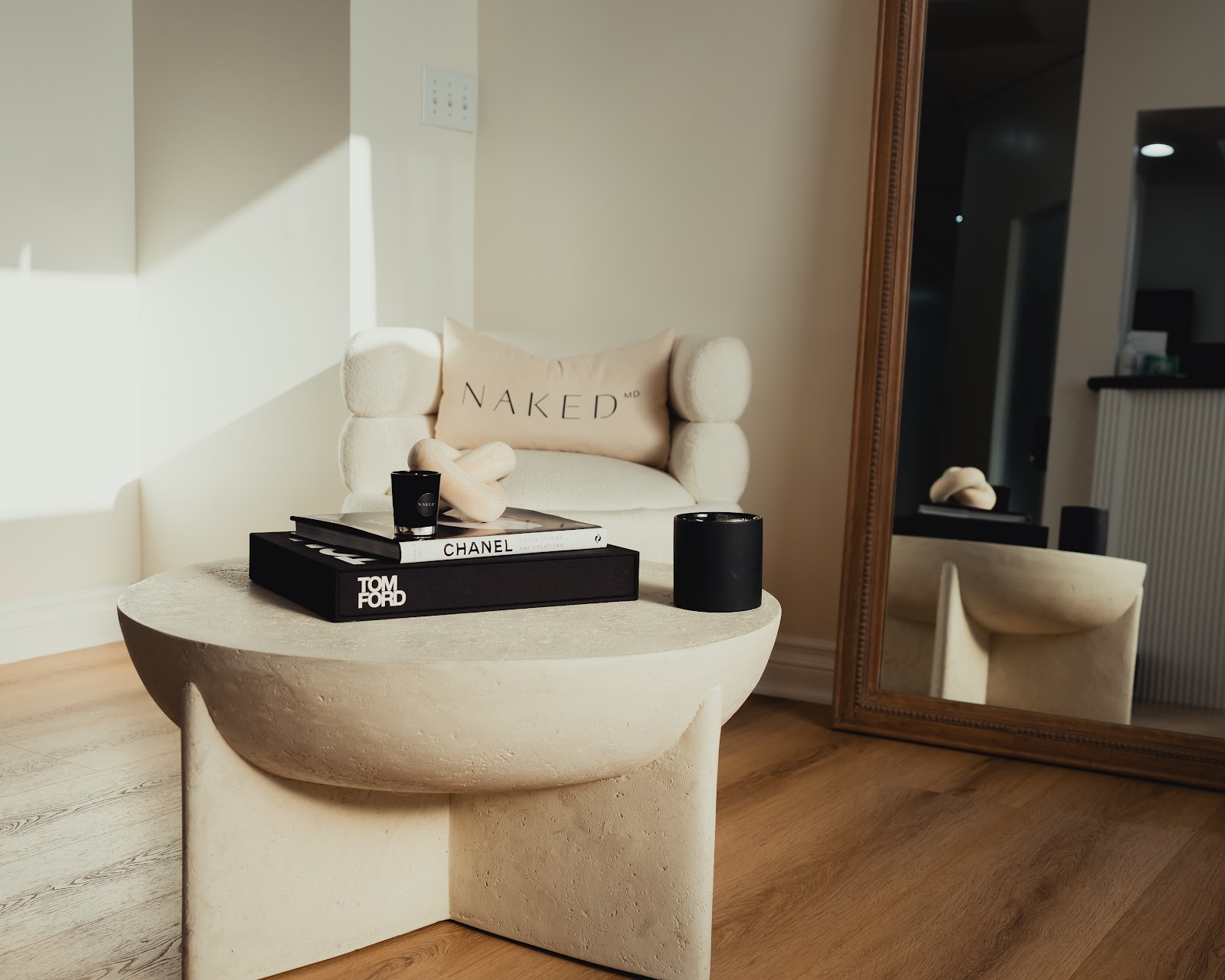 The NakedMD Mission Viejo lobby: a bone boucle chair and round travertine table styled with black Chanel and Tom Ford books and a black candle, beside a tall framed mirror, in soft daylight
