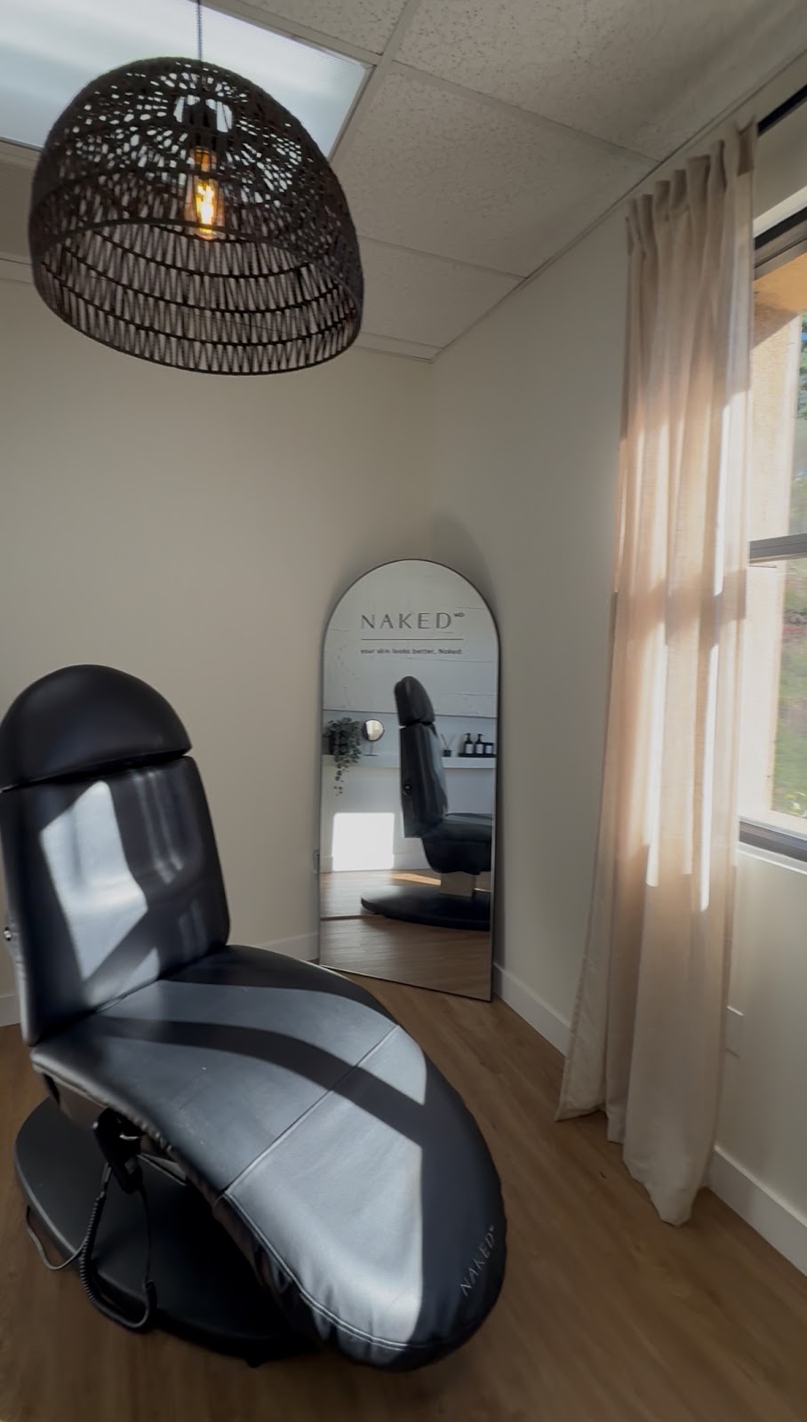 A NakedMD treatment room with a black medical chair, woven pendant, and arch mirror reading your skin looks better, Naked