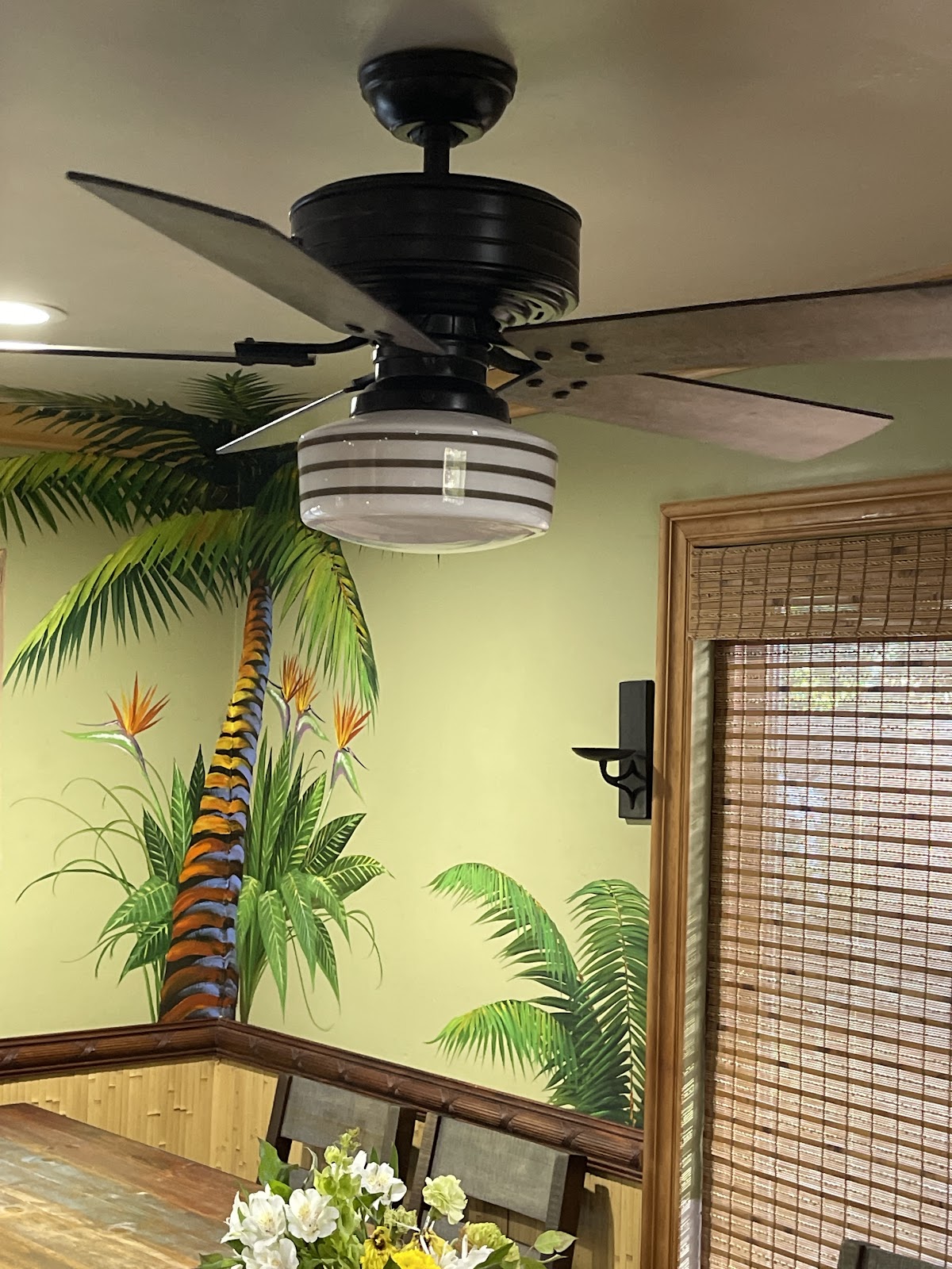 Black ceiling fan with a schoolhouse light installed by O'Hagan Electric over a tropical-mural dining room