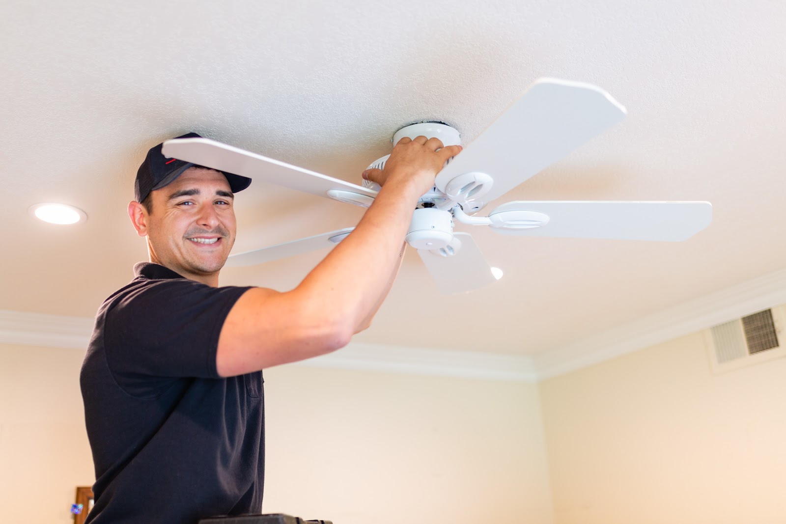 O'Hagan Electric technician installing a white ceiling fan, smiling on a ladder