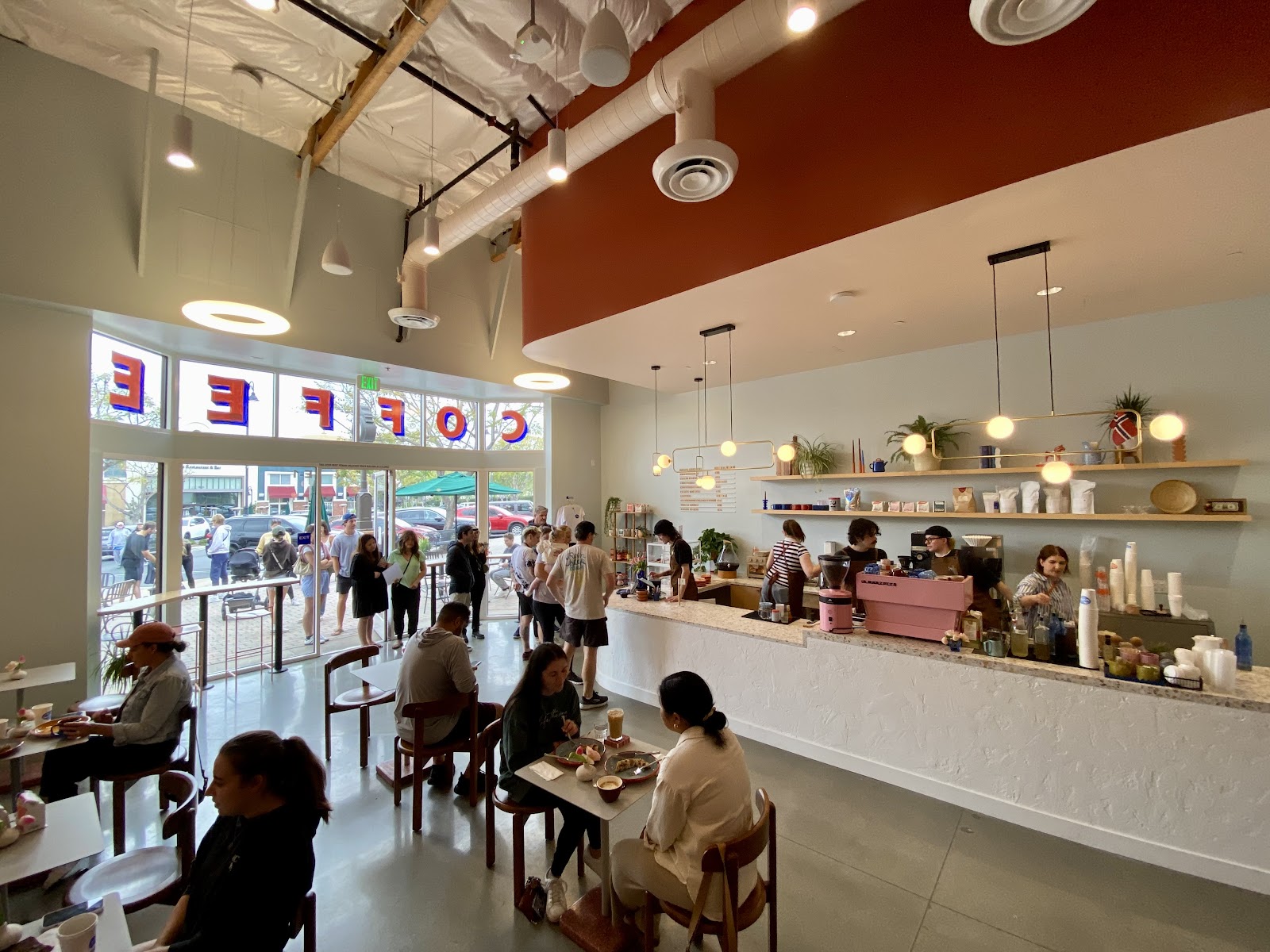 The bright, plant-filled interior of Oslo Coffee Co. with a pink espresso machine on the white counter and customers seated at wood tables