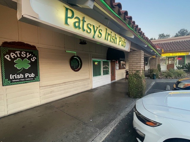 The exterior of Patsy's Irish Pub at dusk, the green channel-letter sign and shamrock logo lit over the plaza