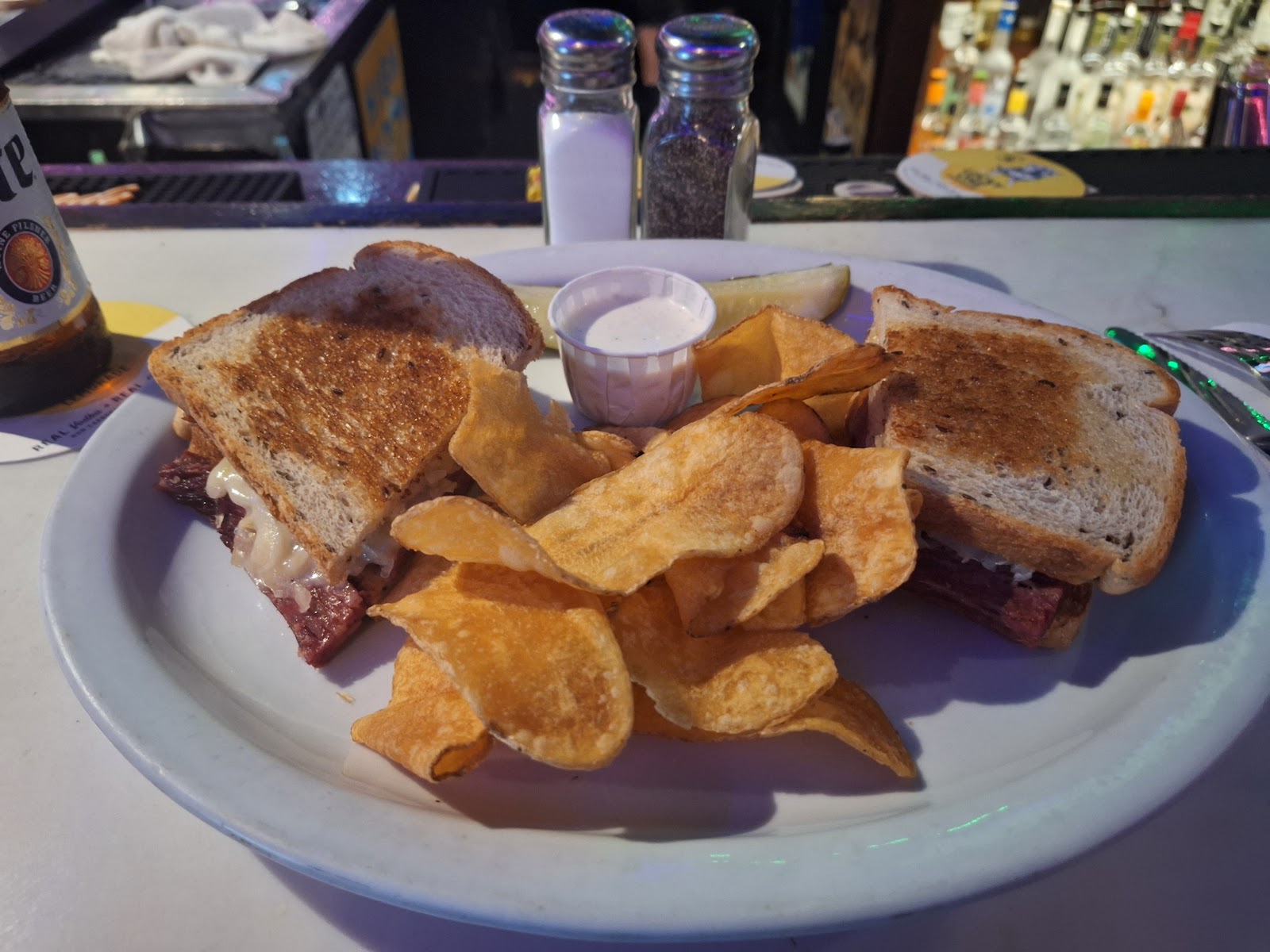A toasted Reuben sandwich on marbled rye with kettle chips and a pickle, a beer bottle beside it
