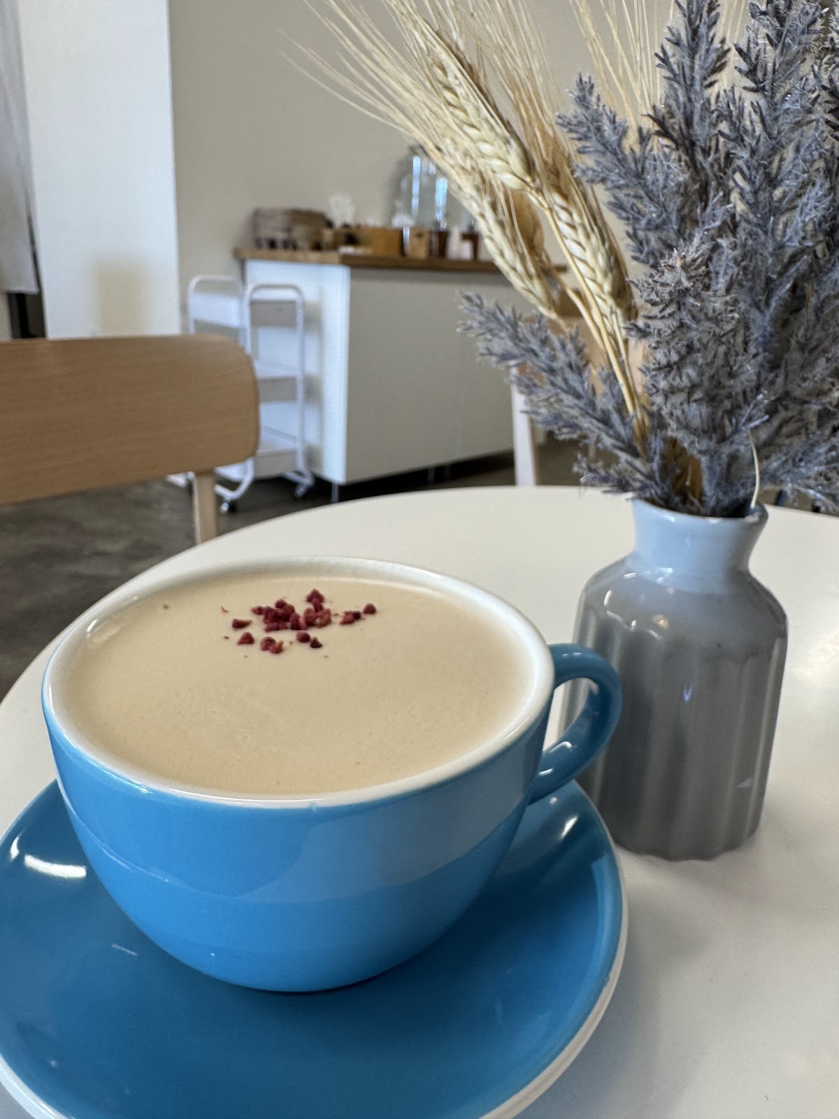 A Vanilla RAF at Redshift, espresso whipped smooth with cream and topped with crushed freeze-dried raspberry in a blue cup beside a vase of dried wheat