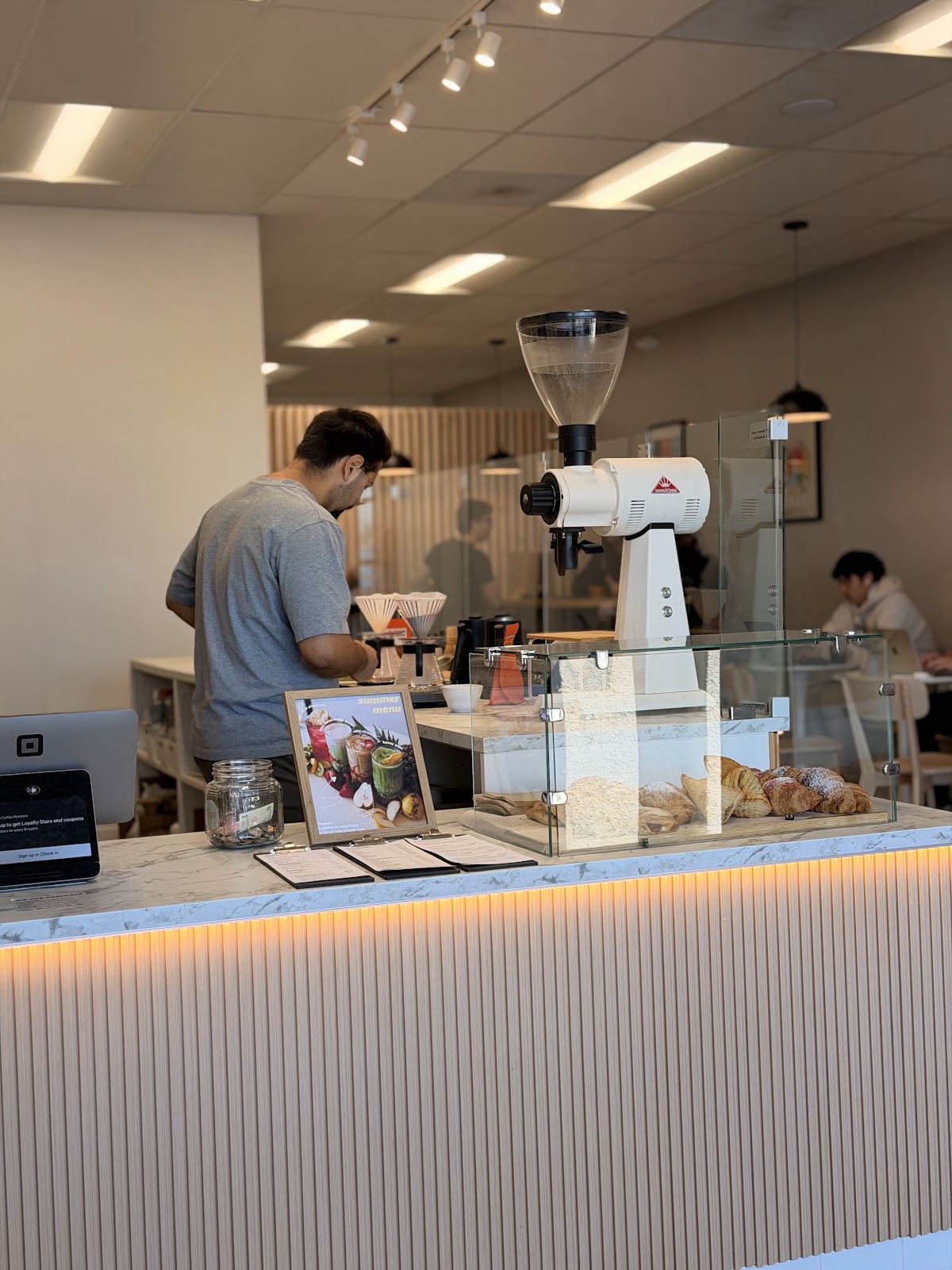 A Redshift barista works pour-over drippers behind the marble counter, a white conical coffee grinder and a glass pastry case of croissants beside the warm under-counter light