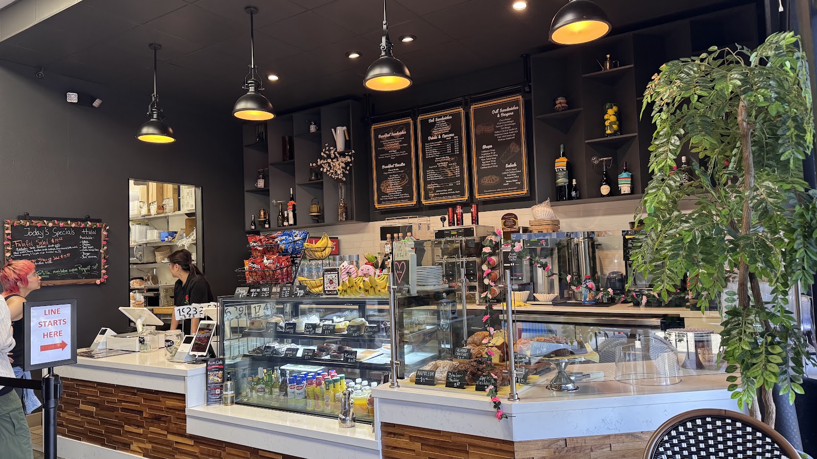 The warm interior of Rose Cafe with chalkboard menu boards, a pastry case, pendant lights, and a large plant