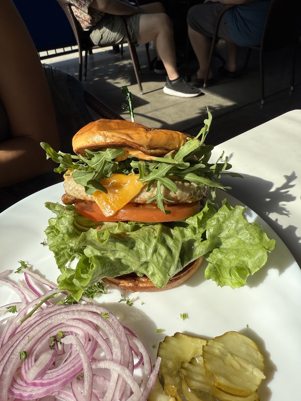 A grilled chicken sandwich on a toasted brioche bun with arugula, tomato, and lettuce, served with pickles and sliced red onion at Rose Cafe