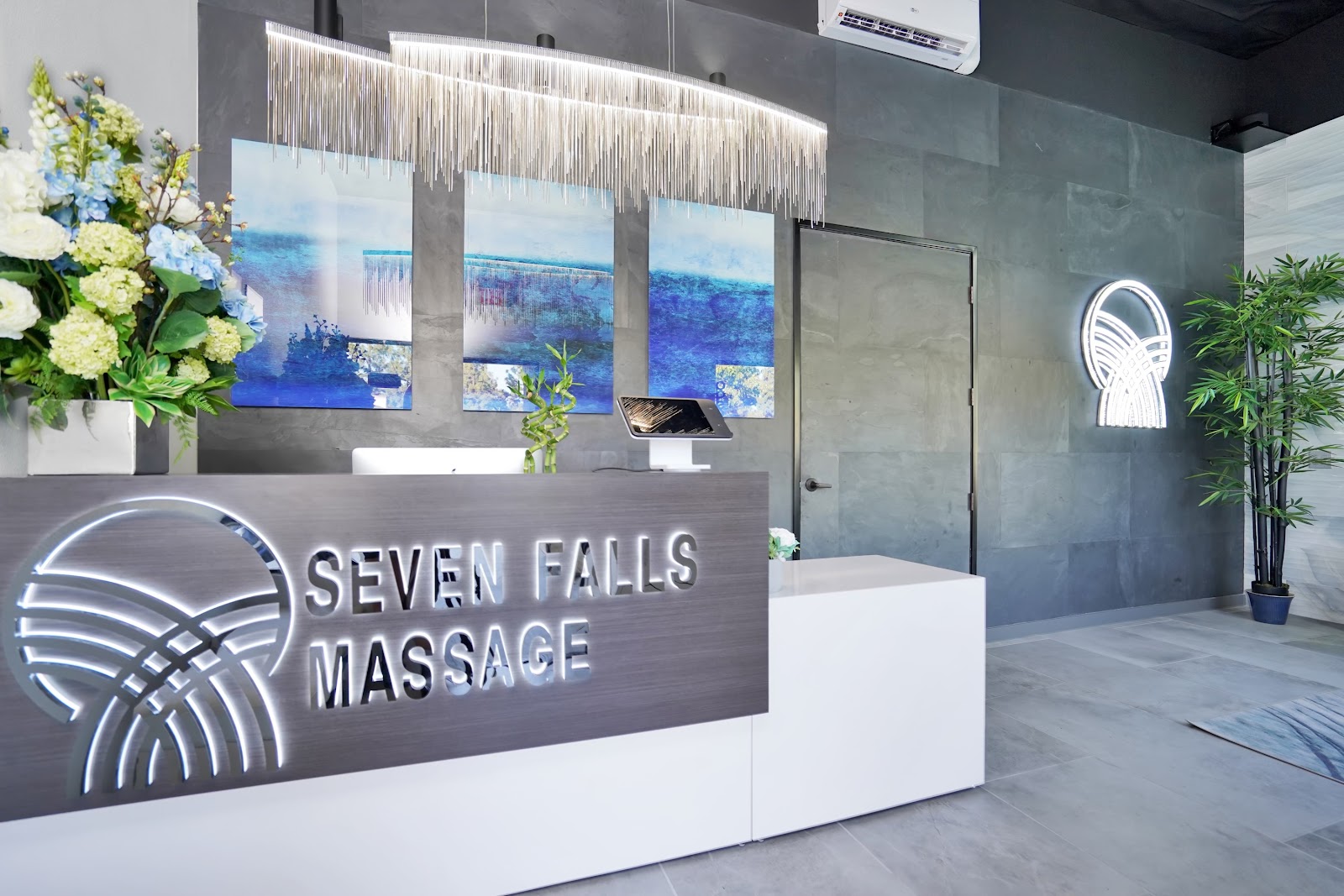 The Seven Falls Massage reception: a slate gray accent wall, a crystal chandelier above the desk, and three abstract blue-water canvases behind the branded sign