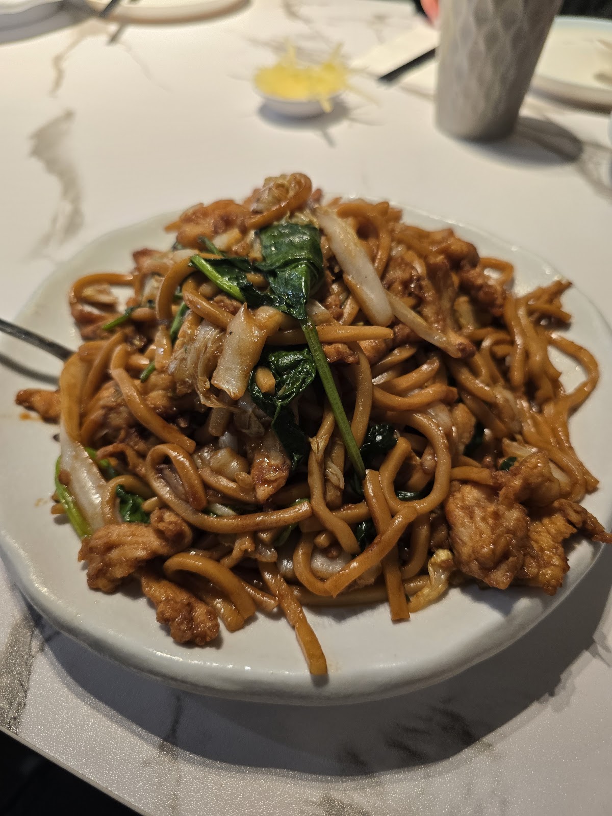 A plate of chicken chow mein with greens and noodles on white marble