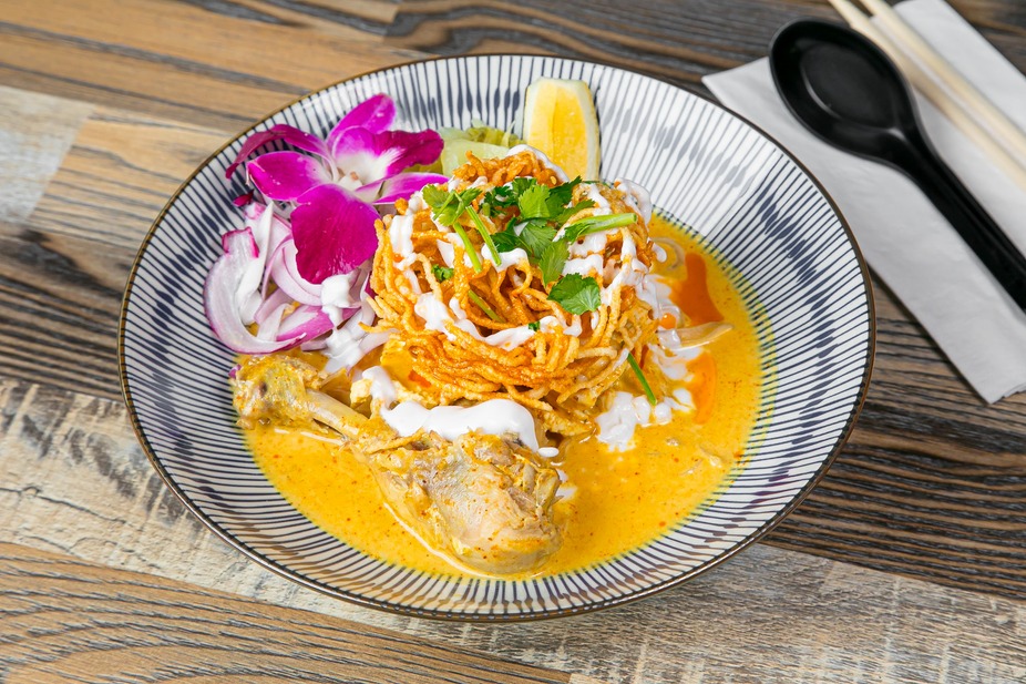 A Thai curry noodle bowl at SIAM with crispy noodles, pickled red onion, lime, and an orchid garnish in golden coconut curry