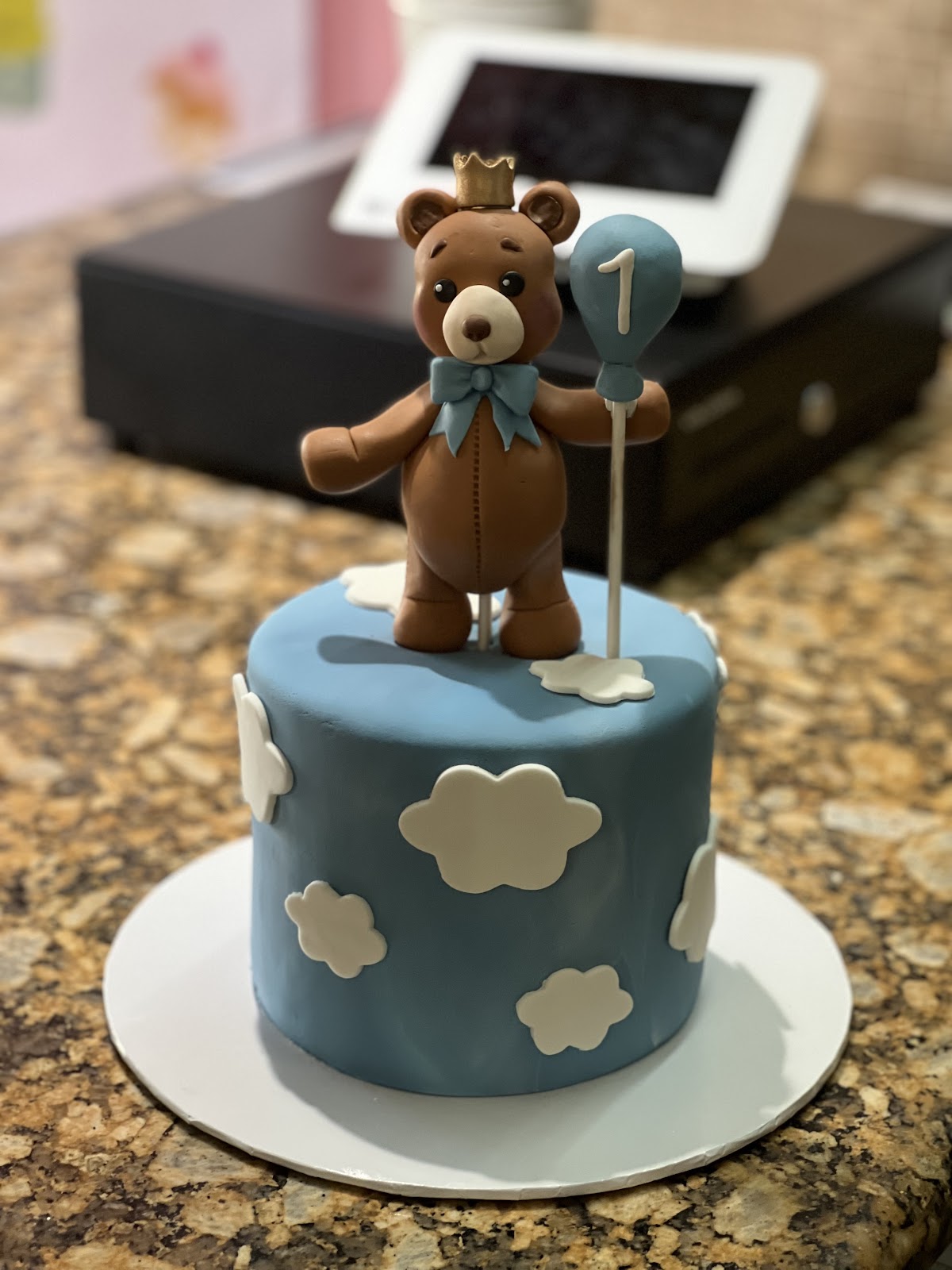 A sky-blue first birthday cake with a hand-sculpted crowned teddy bear, balloon and fondant clouds
