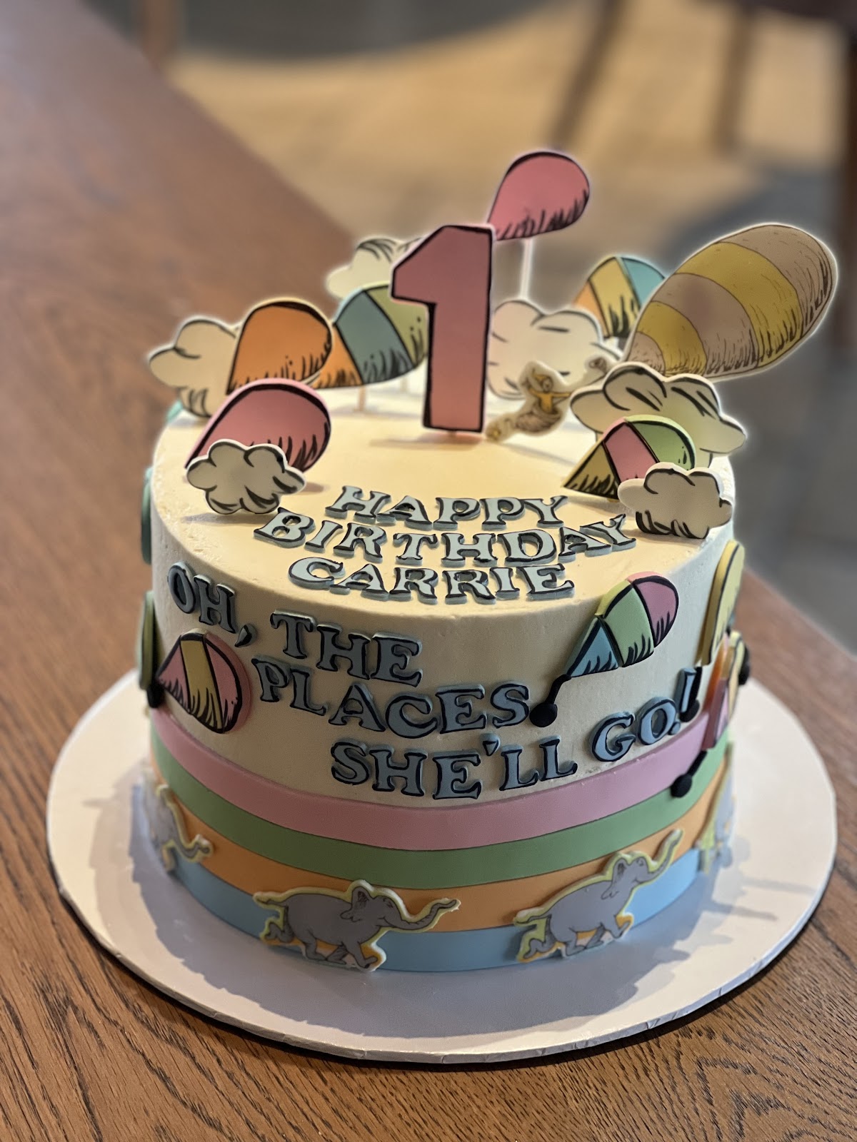 A pastel storybook-themed first birthday cake reading Happy Birthday Carrie, Oh the places she'll go