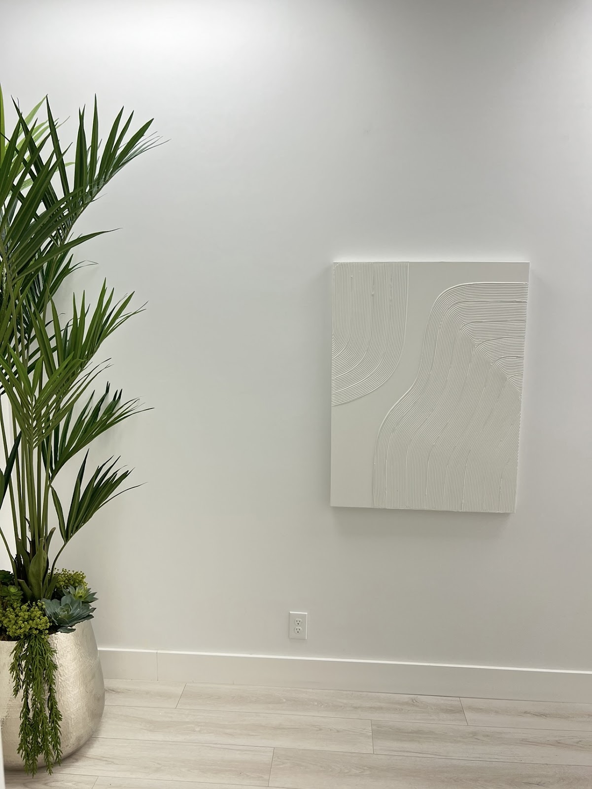 A serene corner of Swan Med Spa: a tall kentia palm beside a white wave-textured plaster art panel on a pale wall