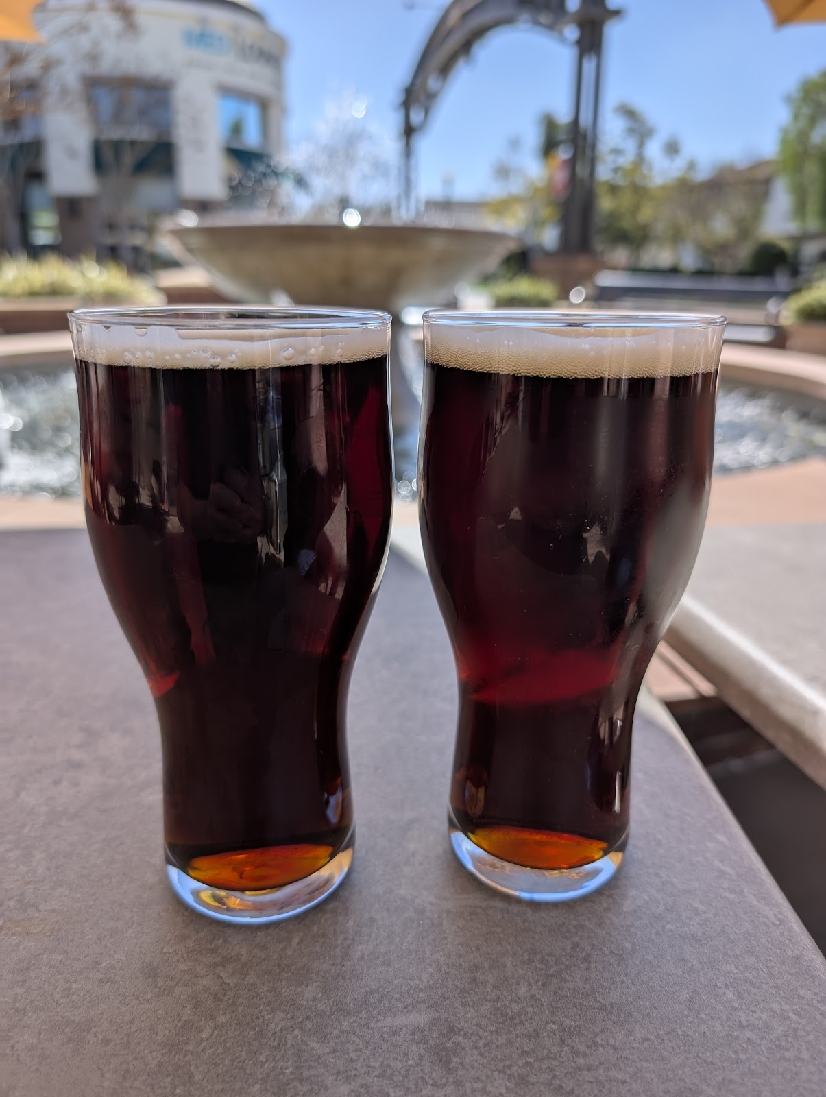 Two dark ales at Ten Mile Brewing glowing amber at the base, on the patio with the plaza fountain behind