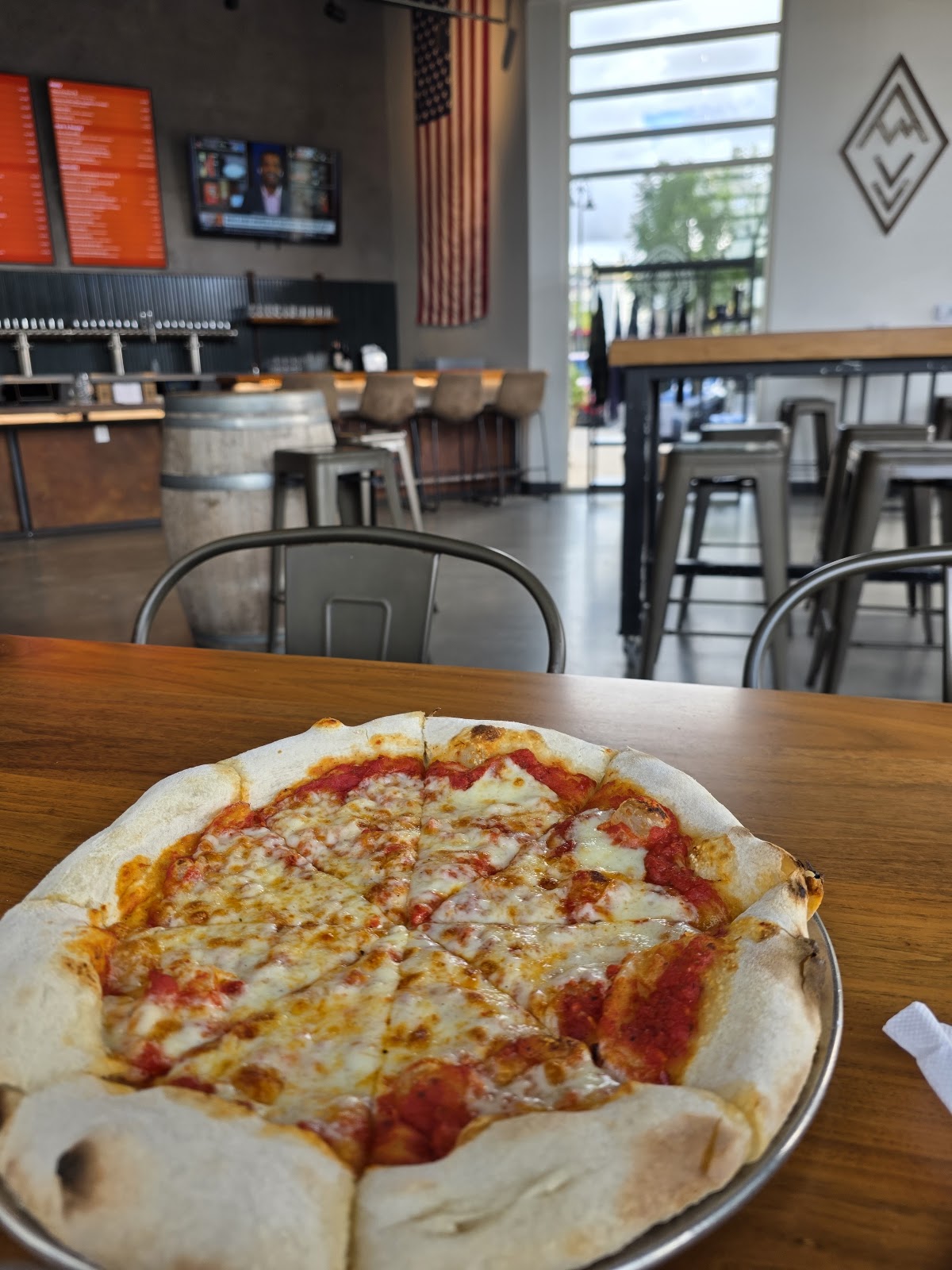 A wood-fired margherita-style pizza on a metal pan at Ten Mile Brewing, the tap wall and a wine barrel behind it