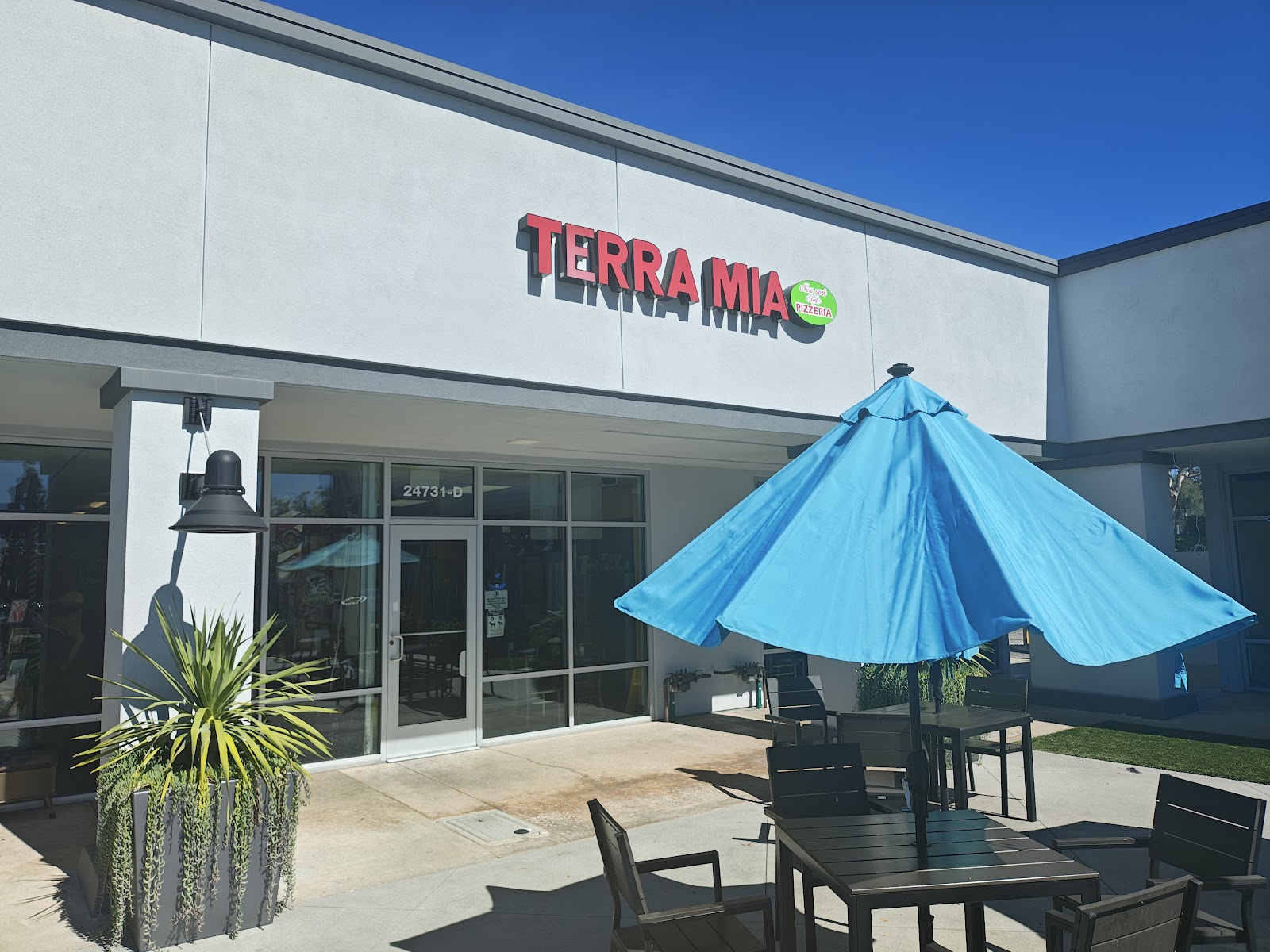The front of Terra Mia Pizzeria on Alicia Parkway, with the red sign, blue umbrella, and outdoor table seating