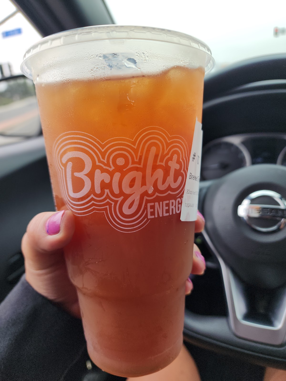 A hand holding a large iced Bright Energy drink in a clear cup with the order ticket, just handed out the drive-thru window into a car