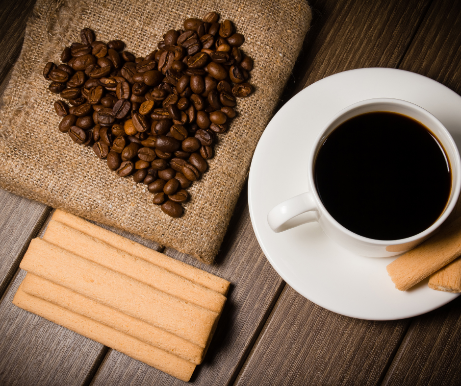 Coffee beans arranged in the shape of a heart on burlap beside a white cup of black coffee