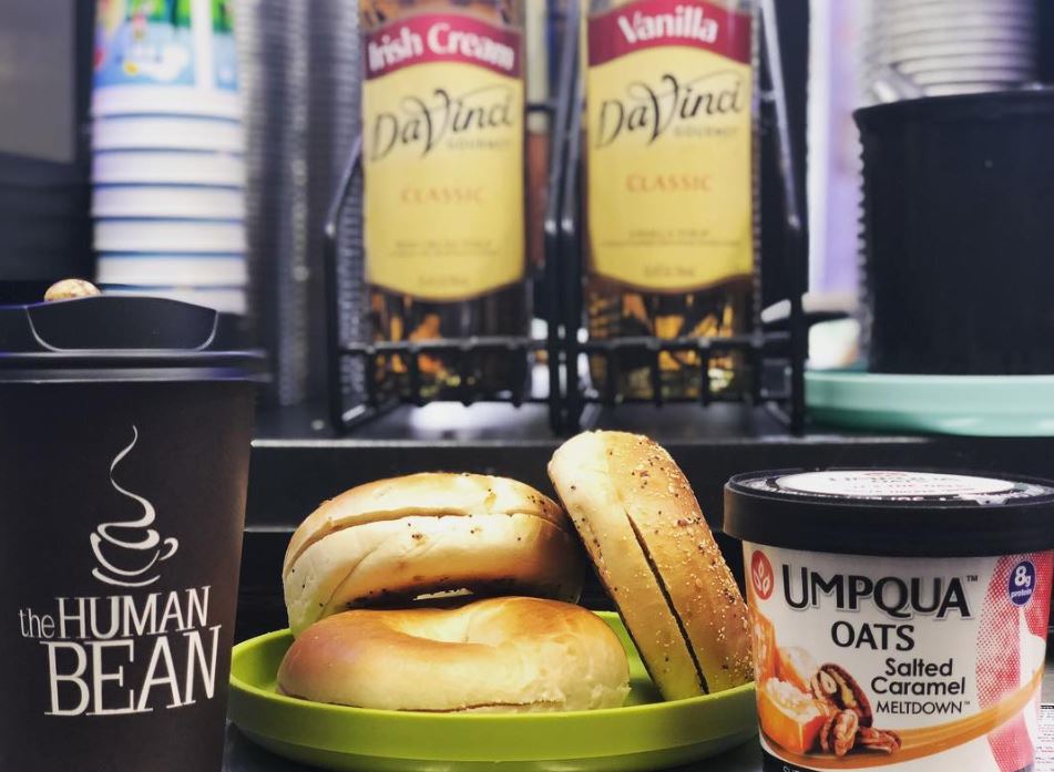 Inside the window: a dark Human Bean cup beside DaVinci Irish Cream and Vanilla syrups, two bagels on a green plate, and Umpqua salted-caramel oats