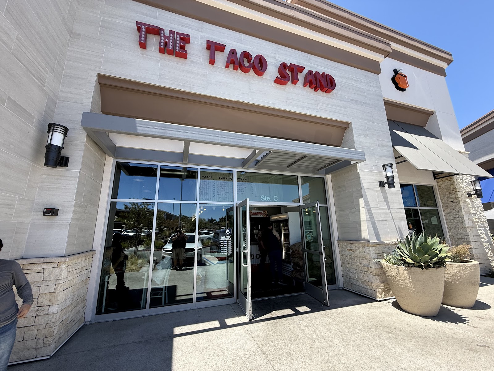The storefront of The Taco Stand in Mission Viejo, bold red THE TACO STAND lettering over the entrance with a chile logo mark and potted agaves by the door
