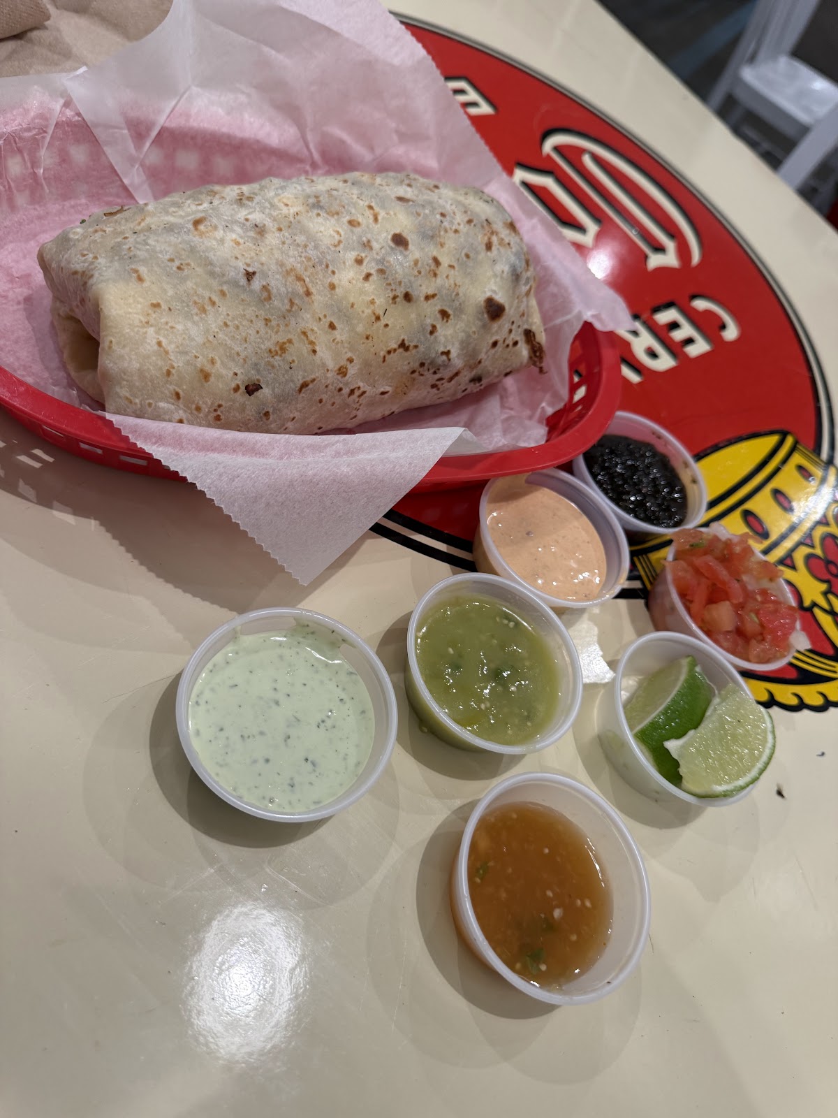 A burrito in a red basket beside an array of house salsas at The Taco Stand: cilantro crema, salsa verde, chipotle, a roja, pico de gallo and lime wedges in small cups